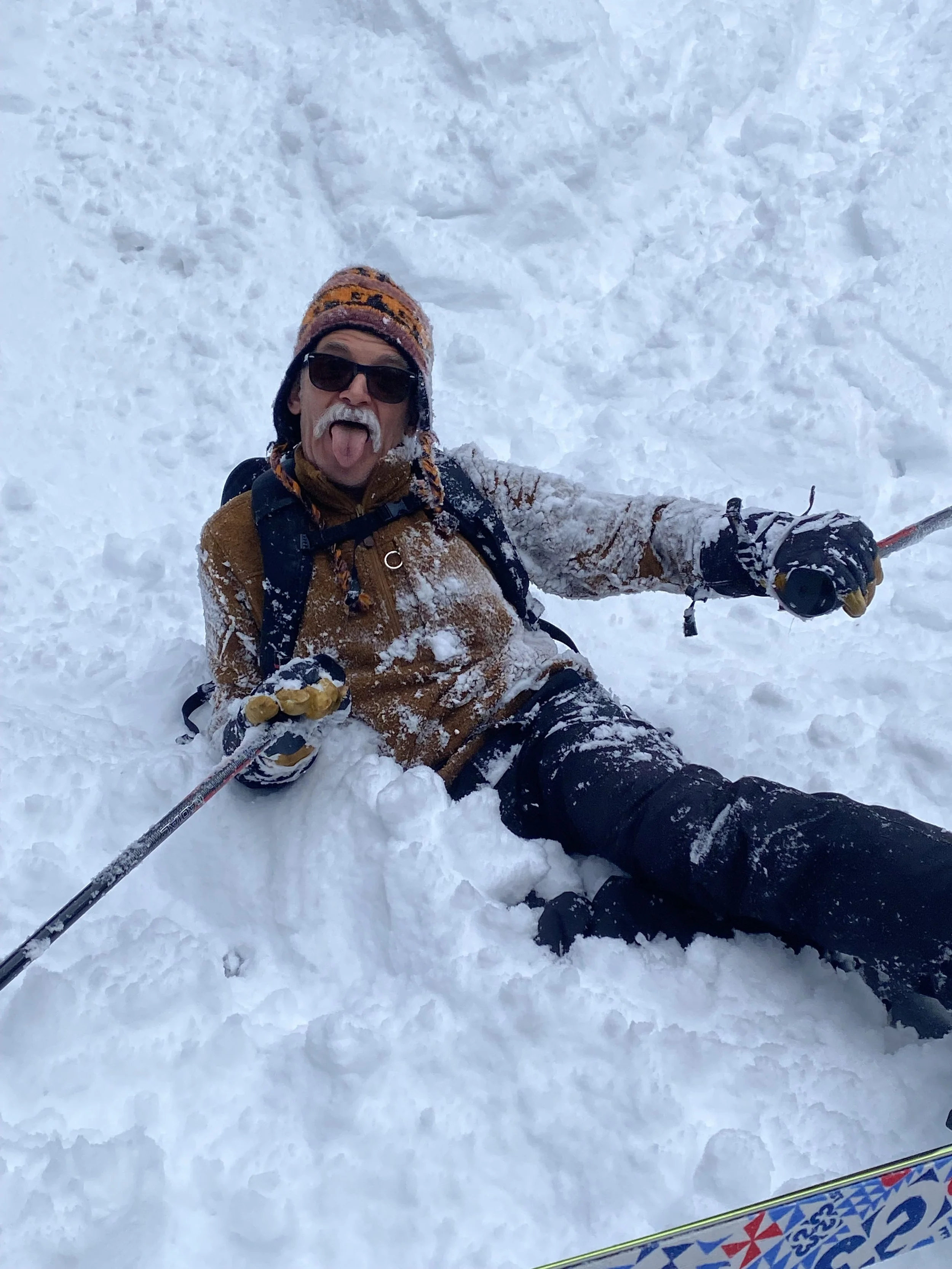 A man with a mustache and sunglasses lying on the snow with a big smile and his tongue out, holding ski poles, wearing a hat, brown jacket, gloves, and black pants, surrounded by snow.
