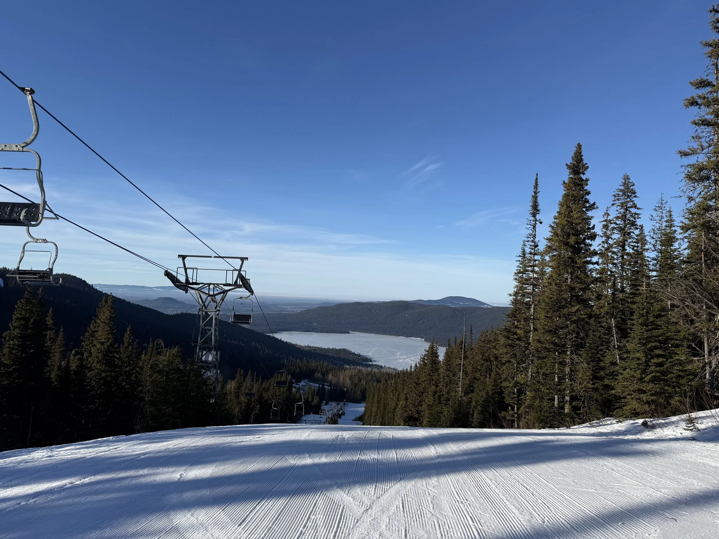 Ski lift ascending over a snowy slope with forested mountains and a lake in the distance under a clear blue sky.