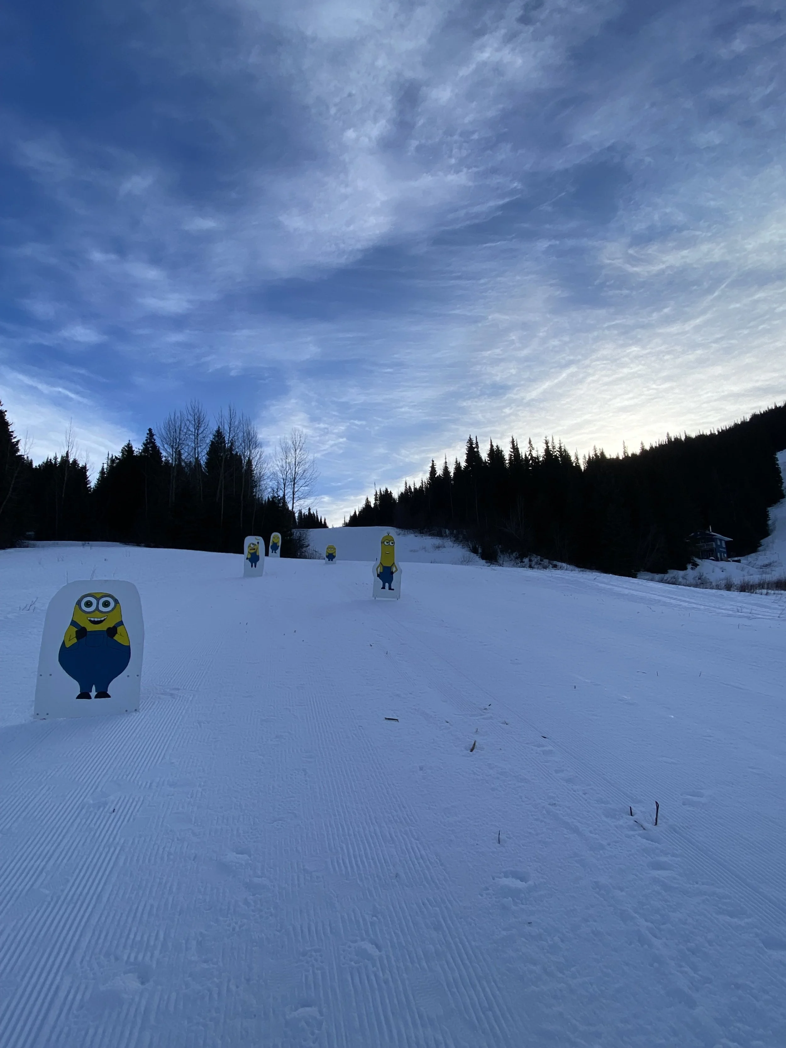 Snow-covered ski slope with cartoon Minions cutouts, surrounded by trees and a blue sky with clouds.