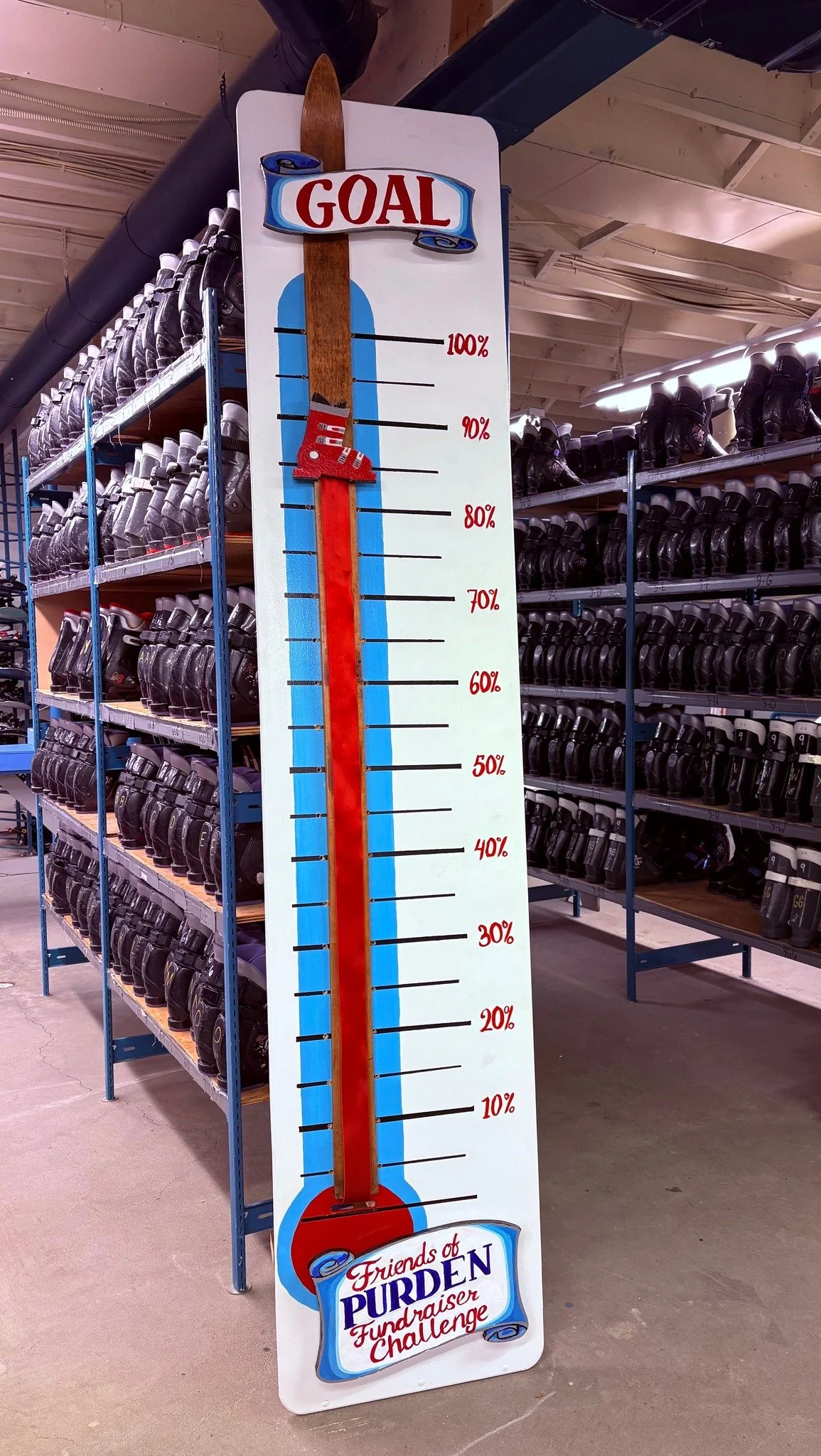 A large goal-themed display with a thermometer-style scale, showing 100% at the top and 0% at the bottom. The display has a wooden paddle with a red sock on it and the logo of 'Friends of PURDEN Gundraiser Challenge' at the bottom.