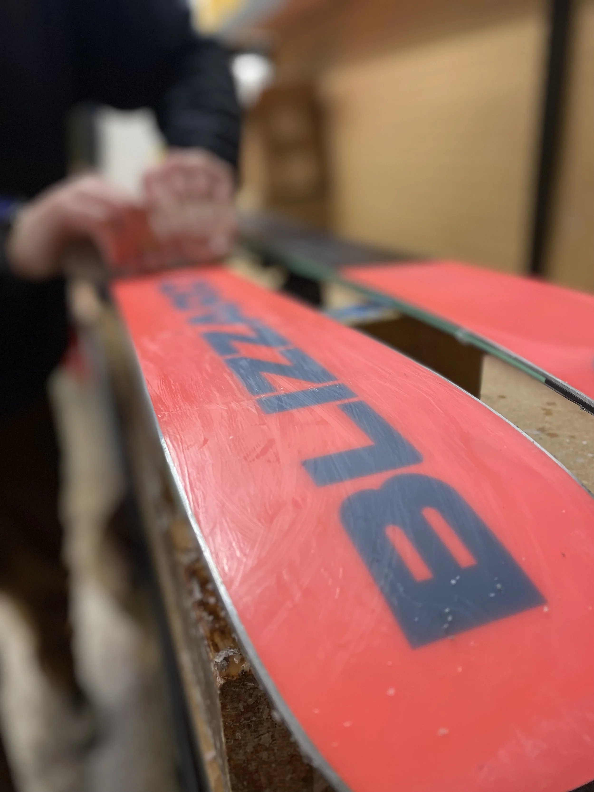 A close-up of a red ski with black lettering, being worked on by a person in a black jacket. The person is holding a tool and appears to be preparing or repairing the snowboard in a workshop.
