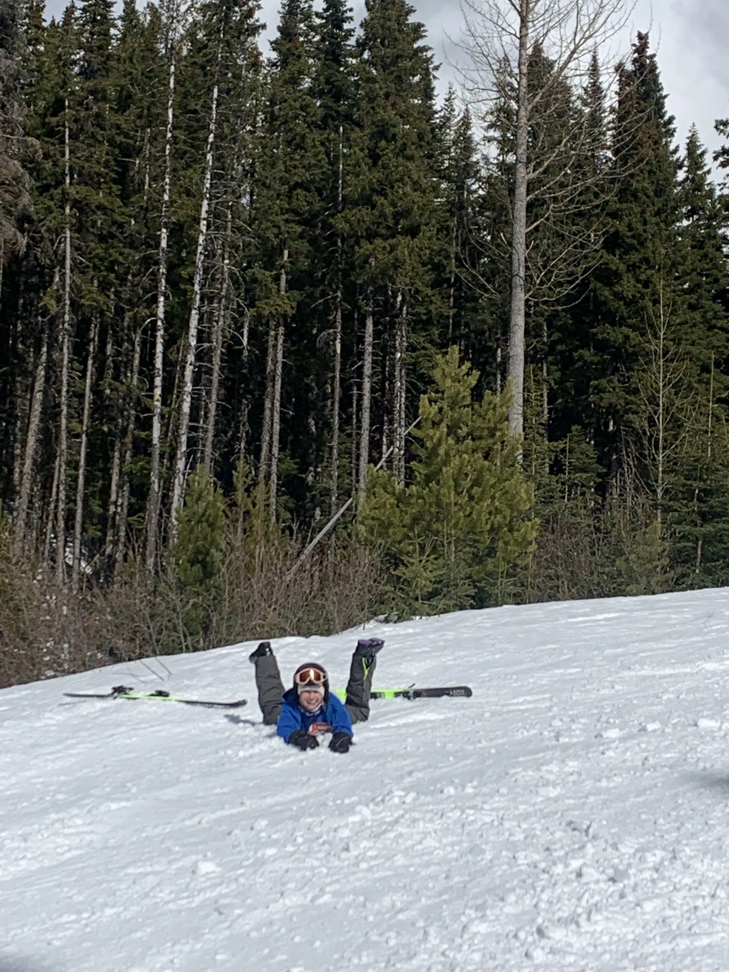 A person lying face down in the snow with skis laid beside, smiling at the camera in a snowy outdoor setting with trees in the background.