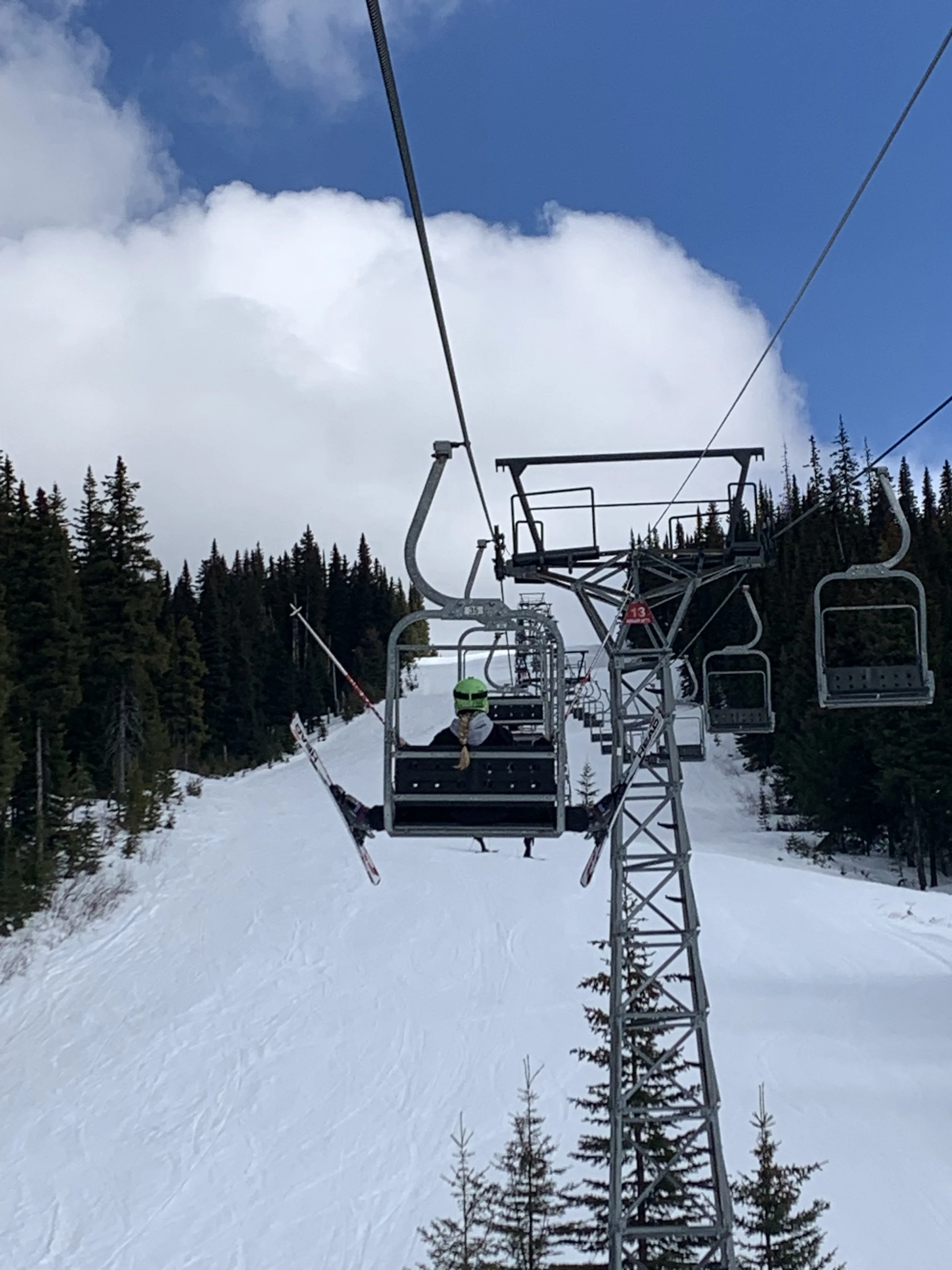 A ski lift with a person wearing a green helmet and black jacket, riding up a snowy mountain surrounded by pine trees under a partly cloudy sky.