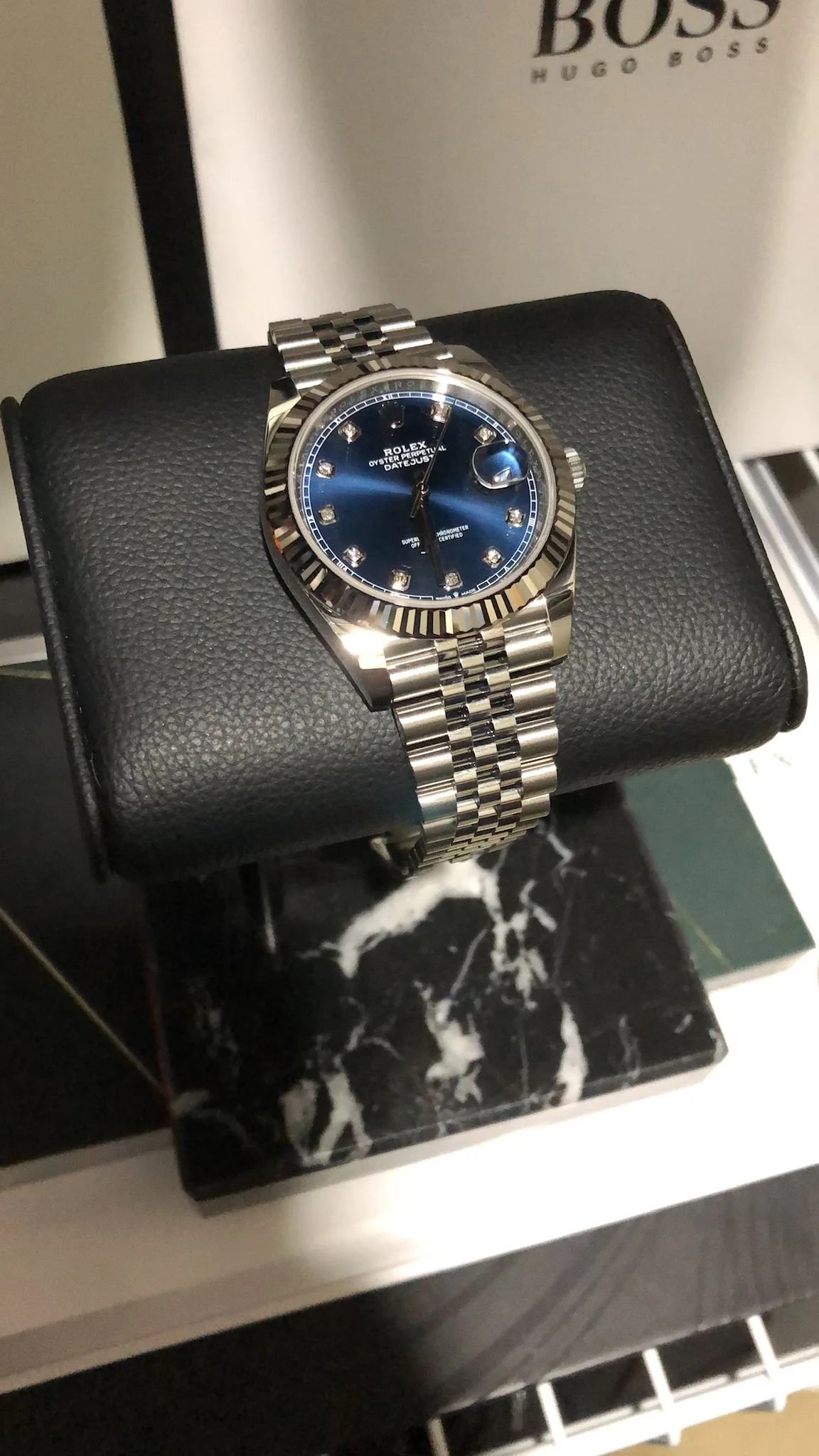 A silver Rolex Oyster Perpetual Datejust wristwatch with a blue dial and diamond hour markers, displayed on a black leather cushion.