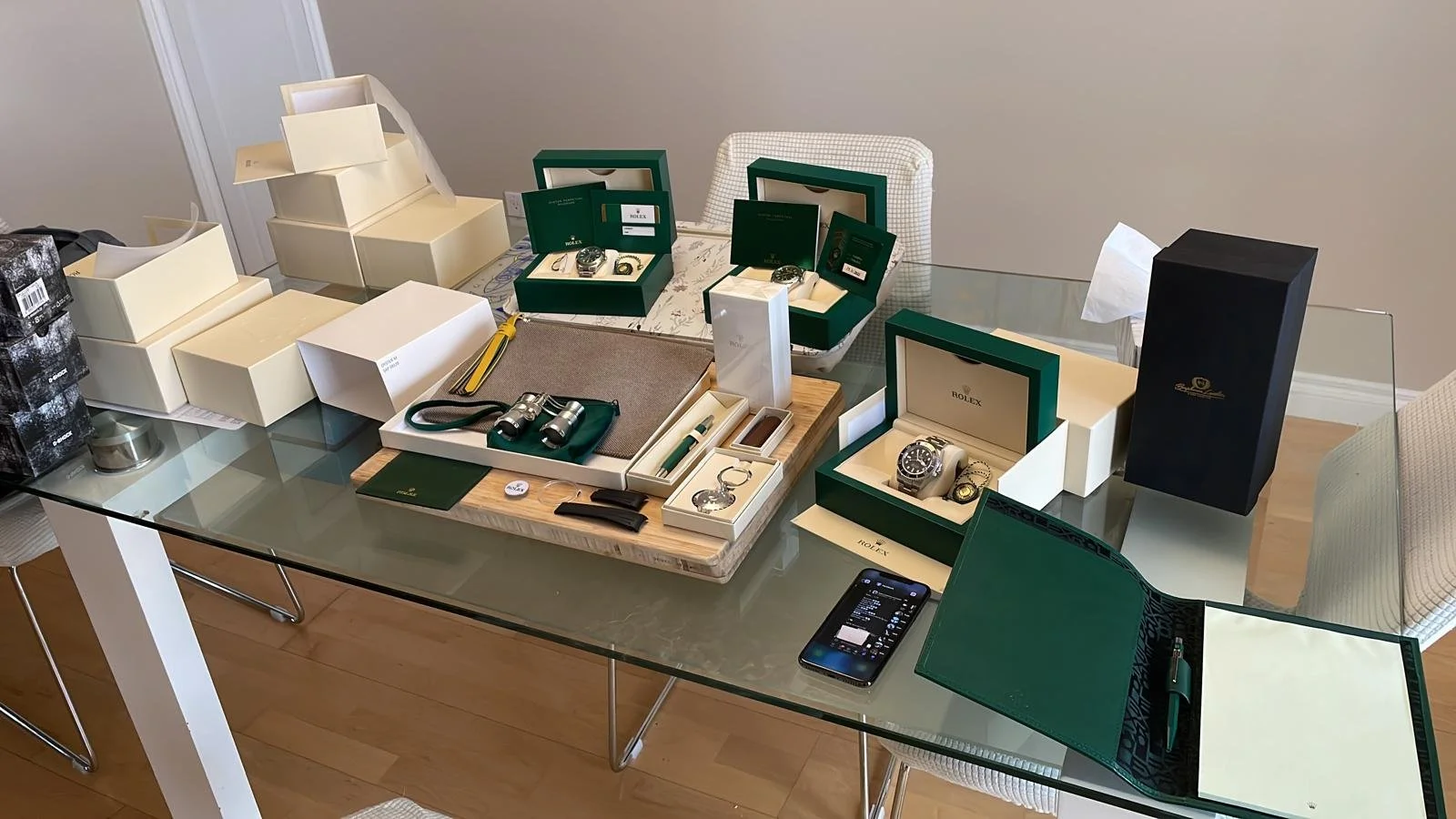 A glass dining table displaying multiple boxed watches, including Rolex, with additional boxes, accessories, and a smartphone, in a room with wooden flooring and beige walls.