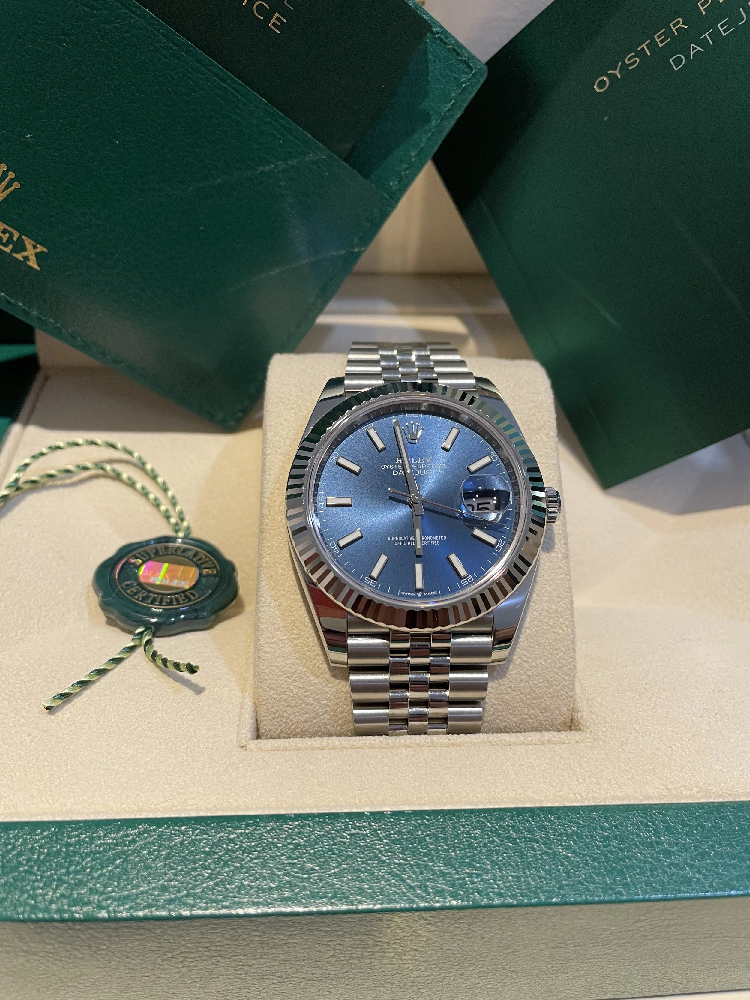 A Rolex Oyster Perpetual Datejust wristwatch with a blue dial, silver hour markers, and a jubilee bracelet, displayed in its original green box with certification tags and packaging.