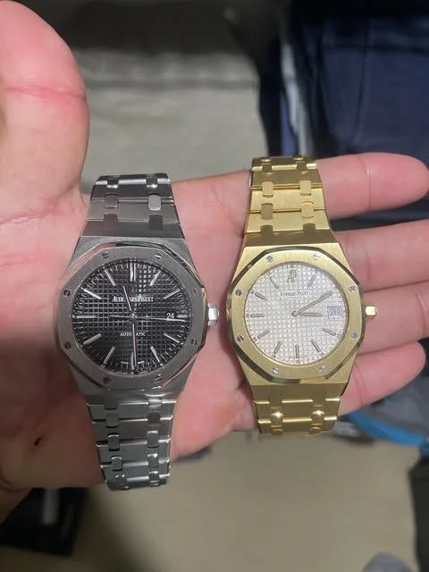 A hand holding two luxury wristwatches, one silver with a black face on the left and one gold with a gold face on the right.