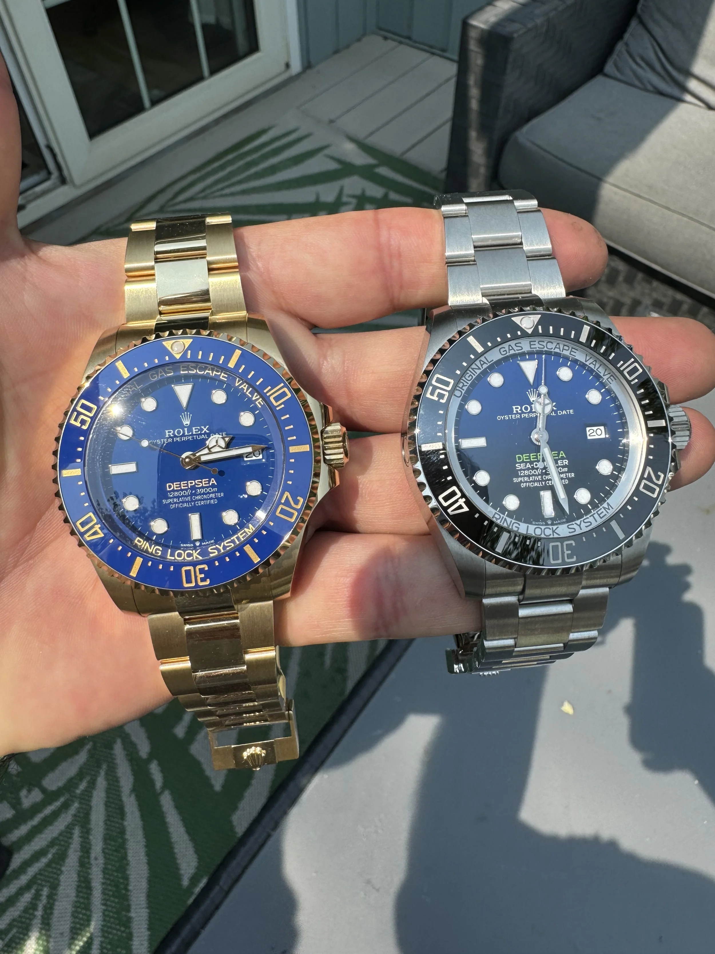 Two Rolex Deepsea watches, one gold with a blue dial and the other silver with a black dial, held in a person's hand outdoors, with patio furniture and a sliding glass door in the background.