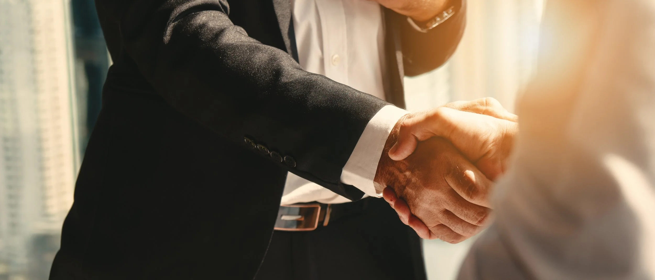 Close-up of two people shaking hands, one wearing a black suit and white shirt, in a professional setting.