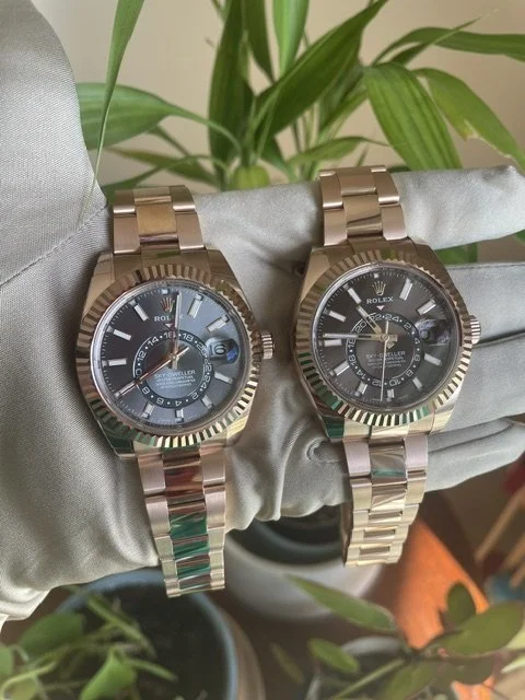 Two gold Rolex watches with black dials and metal bands, placed on a gray fabric surface with green plants in the background.