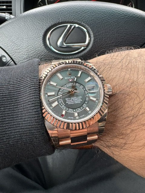 A person's wrist wearing a Rolex Sky-Dweller watch with a green dial, in front of a Lexus steering wheel.