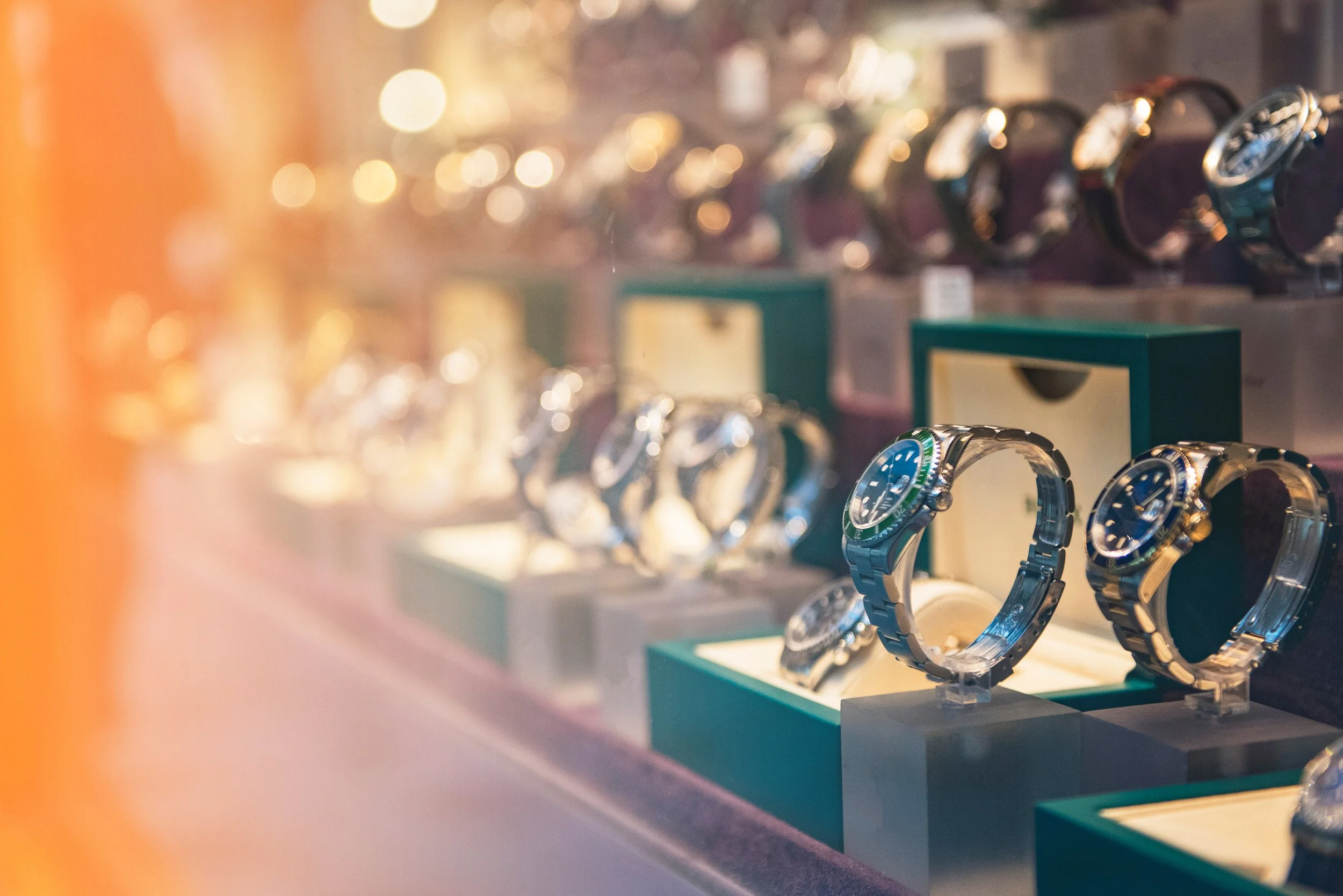 Display case filled with luxury wristwatches, some with metal bands, arranged on stands inside a watch shop with blurred background and warm lighting.