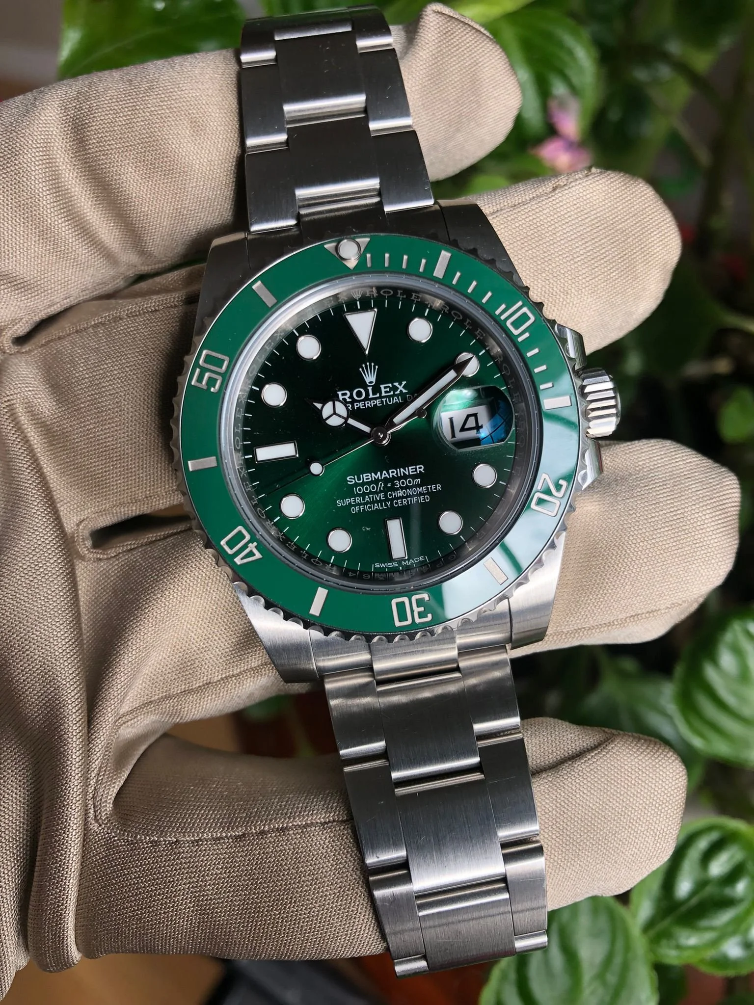 A Rolex Submariner watch with a green bezel and dial, showing the date at 3 o'clock, held by a beige-gloved hand with green foliage in the background.