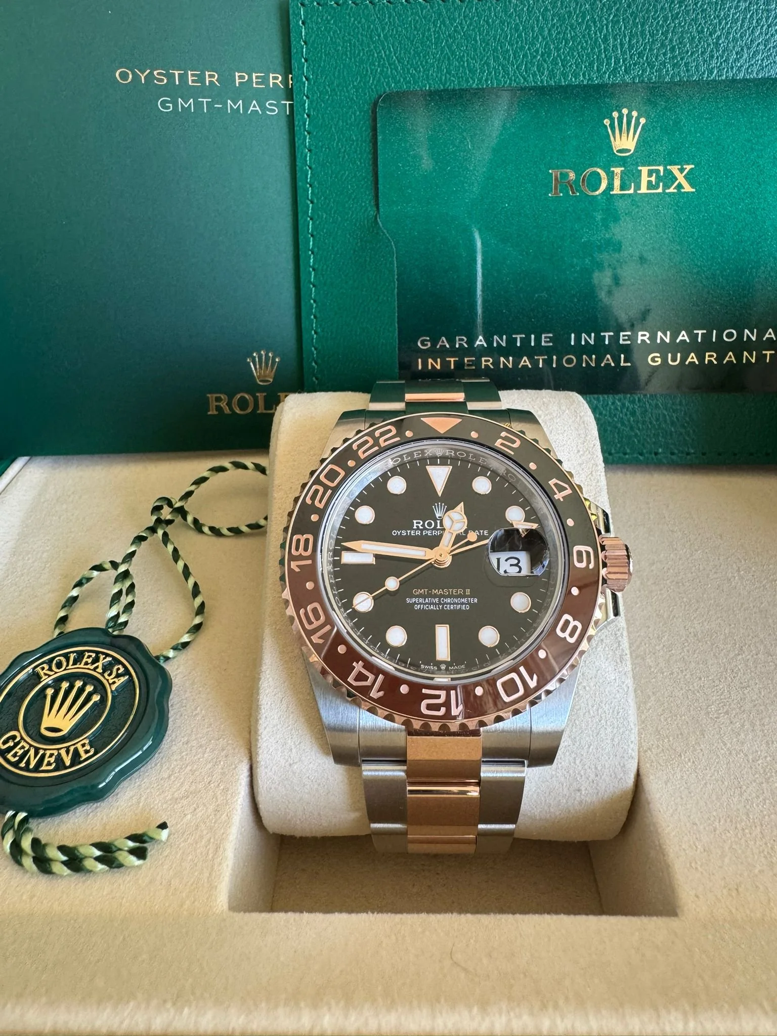 A Rolex GMT-Master II watch with a black dial and two-tone bezel, displayed in a beige box with a green guarantee card and a Rolex tag beside it.