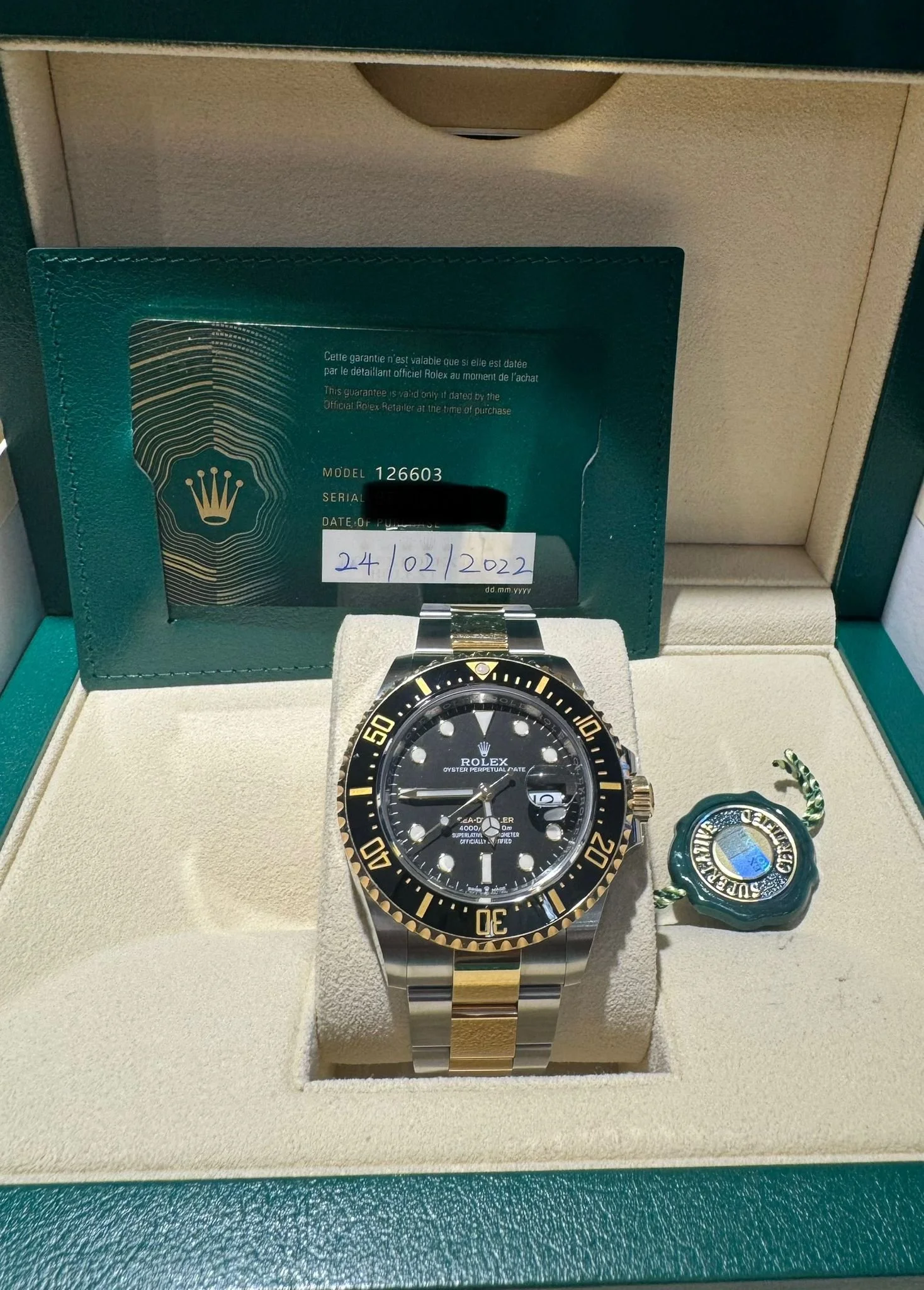 A Rolex Sea-Dweller Deepsea watch with a black dial and a two-tone metal bracelet, displayed in a green and beige watch box along with a green tag and a Rolex warranty card dated 24/02/2022.