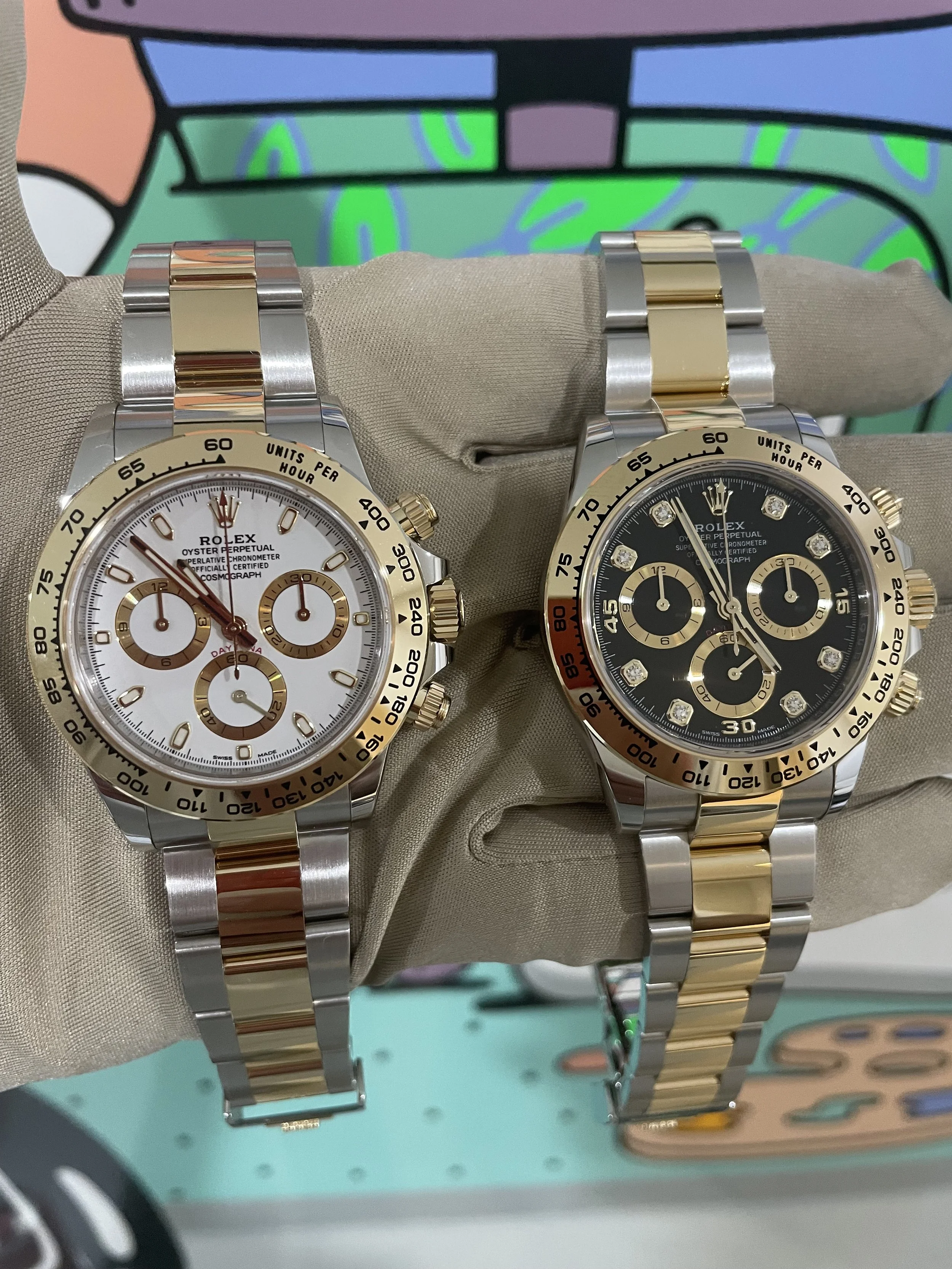 Two Rolex watches with metal bands, one with a white dial and gold accents, the other with a black dial and gold accents, displayed on a cloth holder against a colorful background.