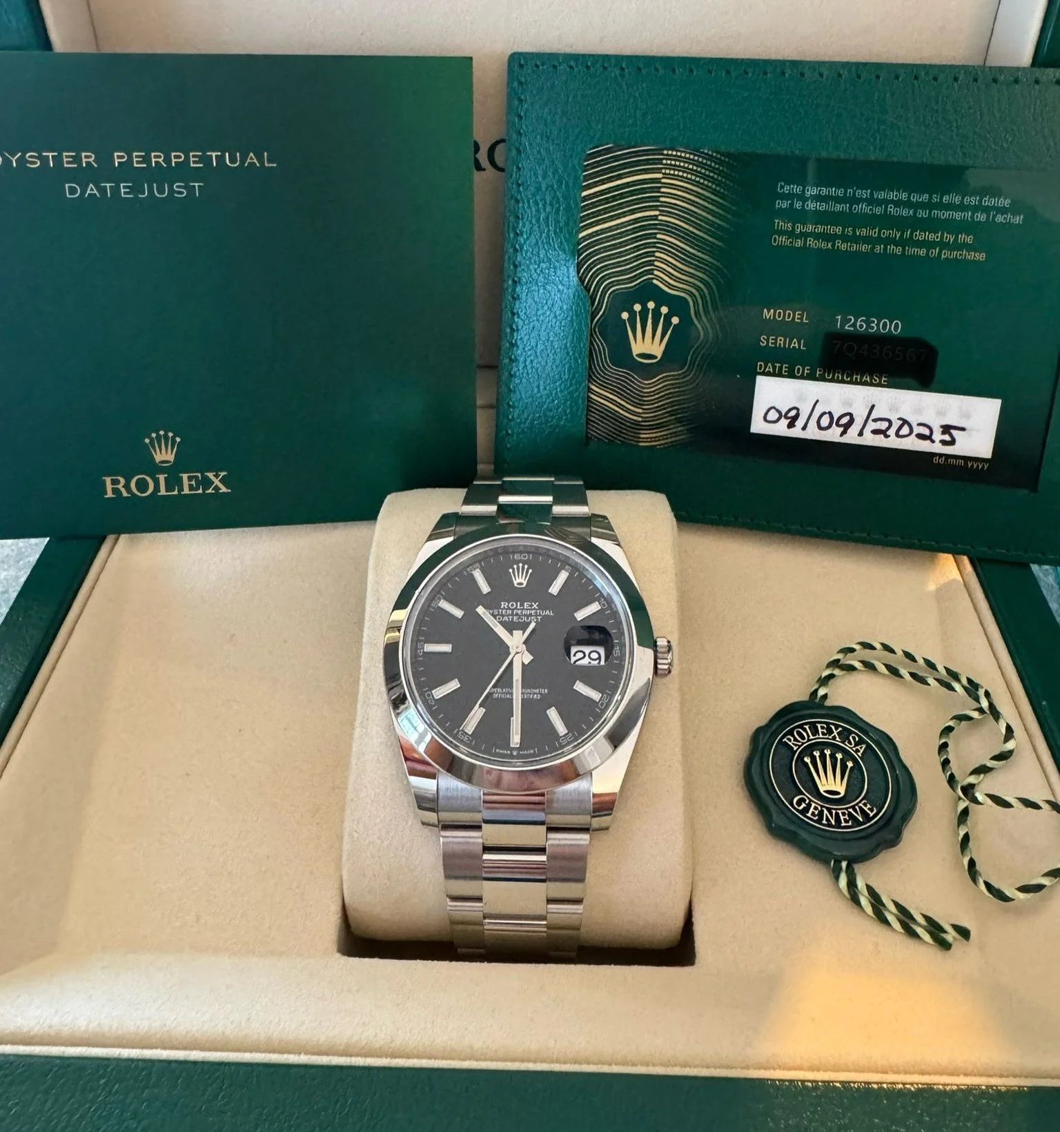 A Rolex Oyster Perpetual Datejust wristwatch with a black dial, silver markers, and a date window, placed on a beige cushion inside its green box. The box contains official documentation, a warranty card, and a green tag with a black and white string.