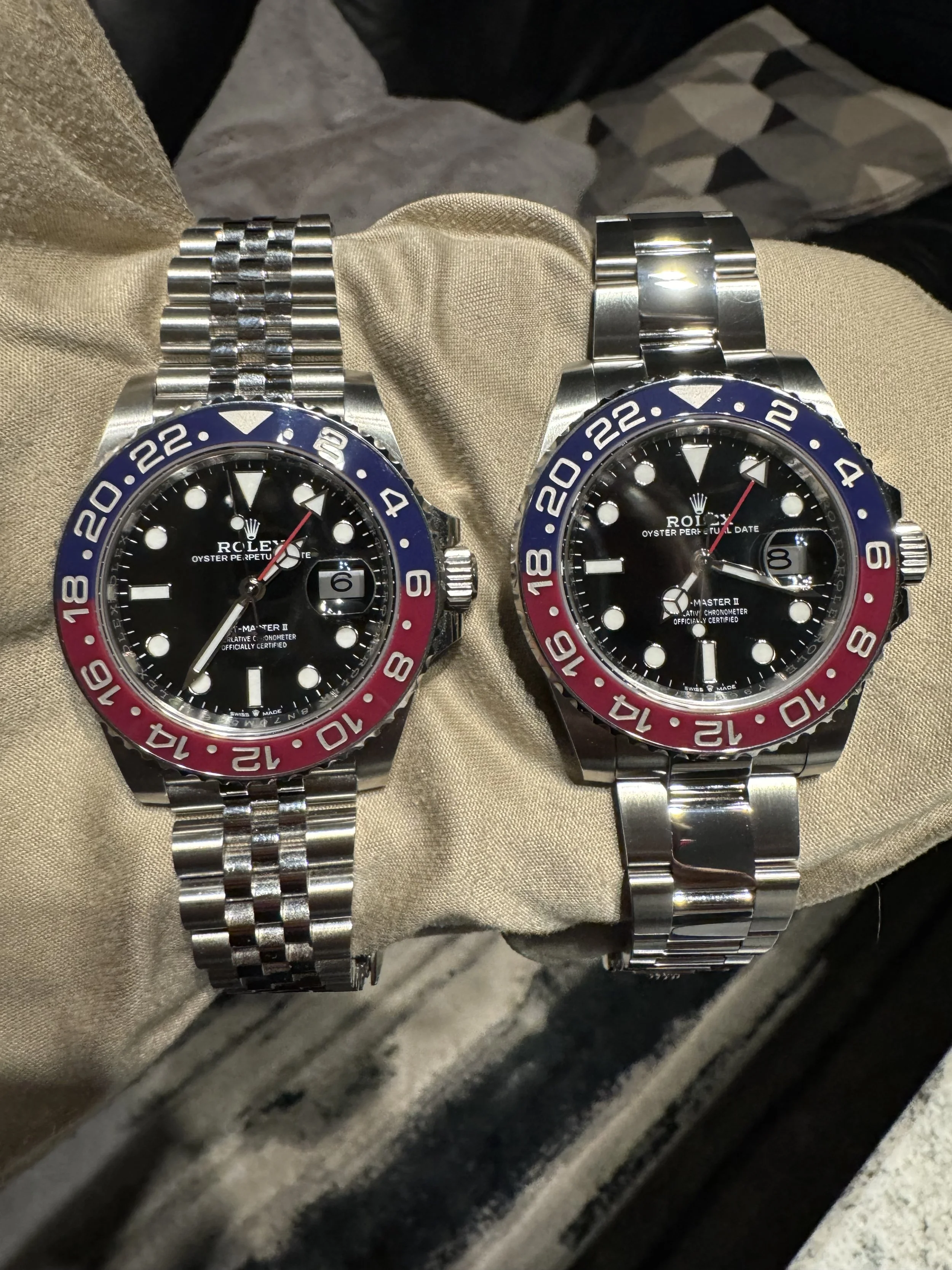 Two Rolex watches on a beige cloth, both with black dials and red and blue bezels. The left watch has a jubilee bracelet, and the right watch has a flat link bracelet.