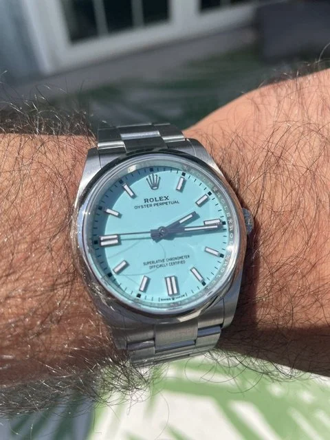 A person's wrist wearing a silver Rolex Oyster Perpetual watch with a light blue dial and silver hour markers, outdoors with a house and green grass in the background.
