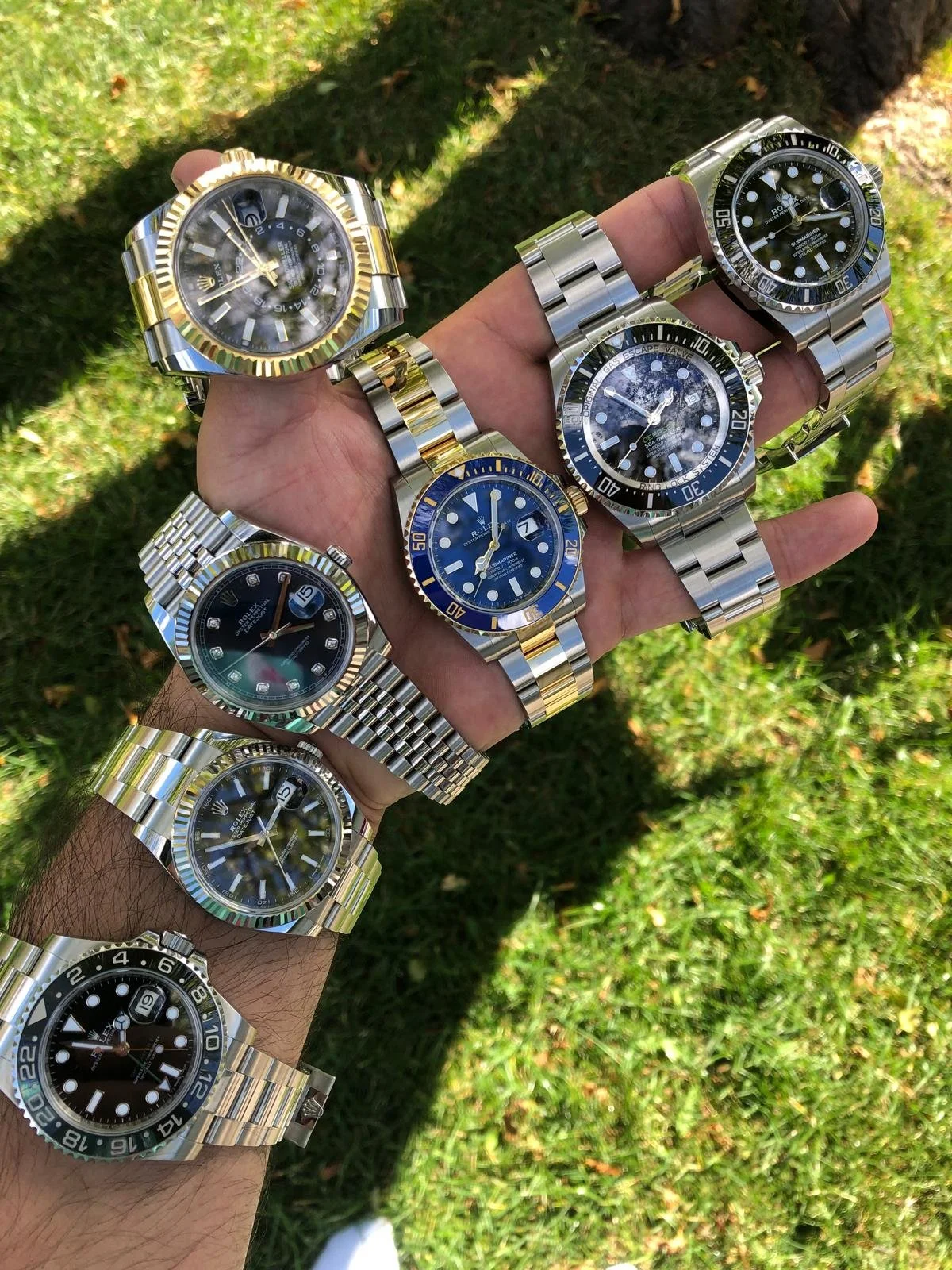 Hand holding multiple luxury Rolex watches with a grassy background.
