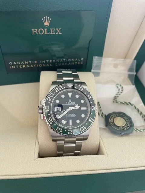 Rolex black and silver wristwatch displayed in an open green Rolex box with a beige cushion, with a green and white Rolex tag and a booklet in the background.