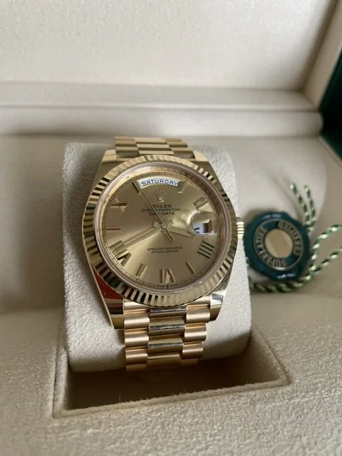 A gold Rolex Day-Date watch with a gold dial, Roman numeral hour markers, and a date window at 3 o'clock, displayed on a soft cushion in a box.