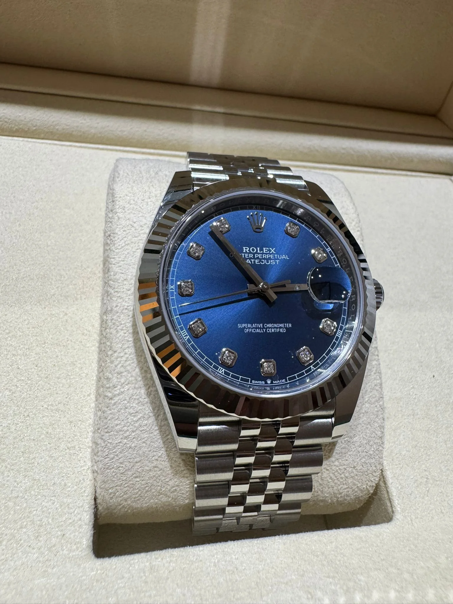 A Rolex Oyster Perpetual Datejust watch with a blue dial, diamond hour markers, a stainless steel bracelet, and a fluted bezel inside a beige watch box.