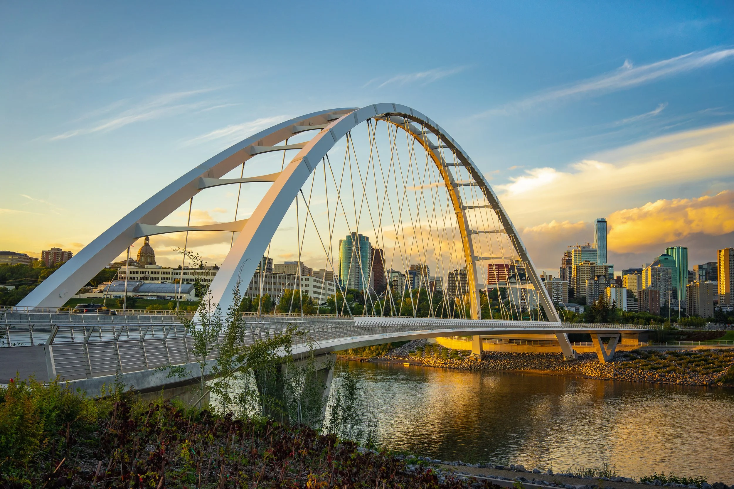 A modern white arched bridge crossing a river with a city skyline featuring tall buildings in the background, under a partly cloudy sky during sunset.