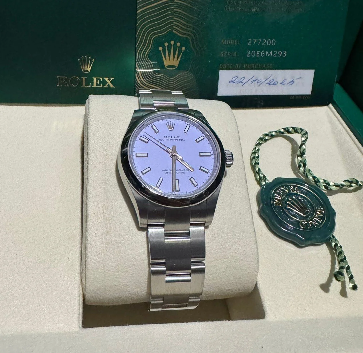 A Rolex wristwatch with a silver band and a white face, displayed in a beige presentation box. There is a green Rolex tag with a black cord beside the watch, and a green Rolex certificate in the background.