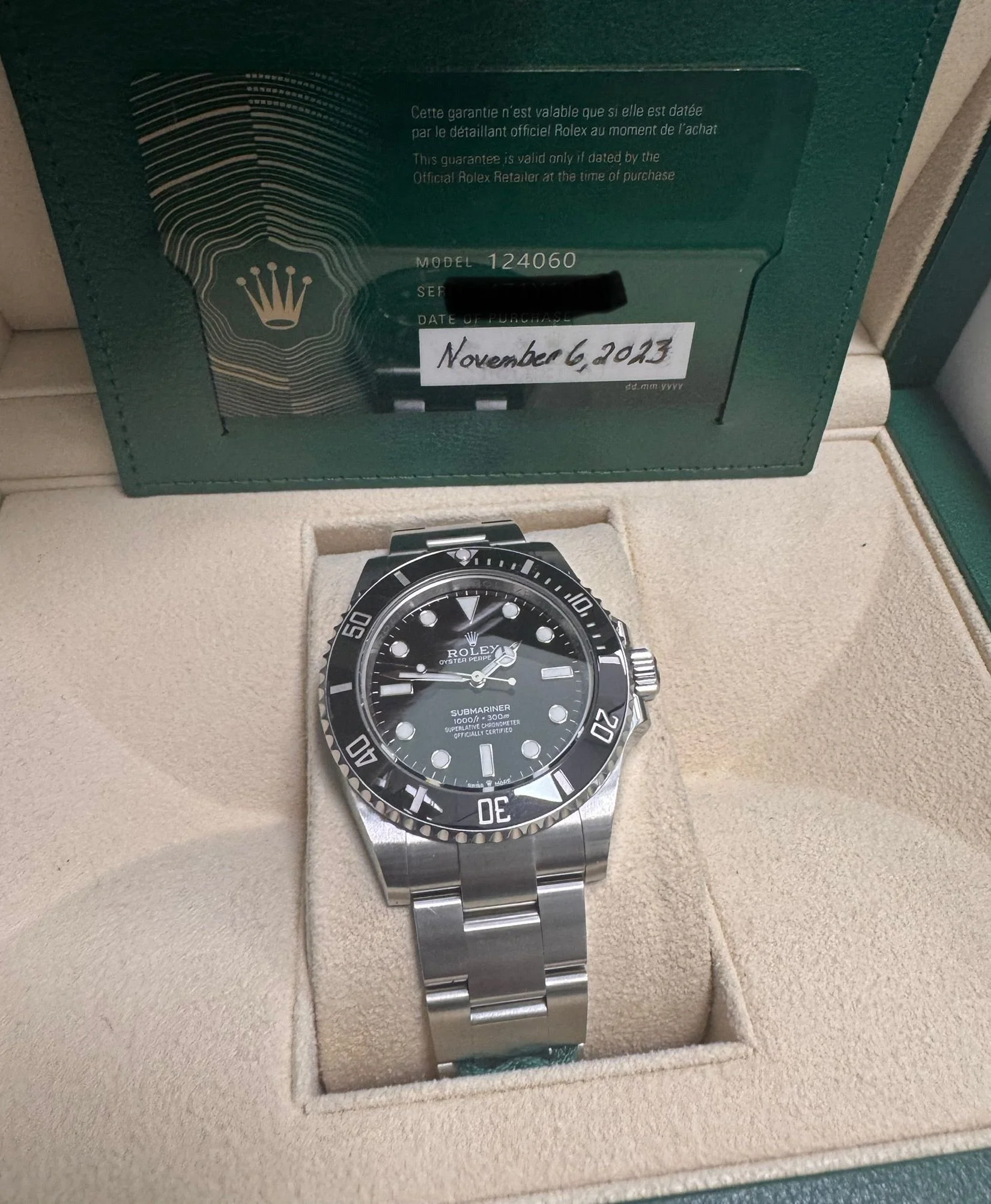 A stainless steel Rolex Submariner watch with a black dial and bezel, resting in a beige watch box, with a Rolex guarantee card in a green holder displayed behind it.