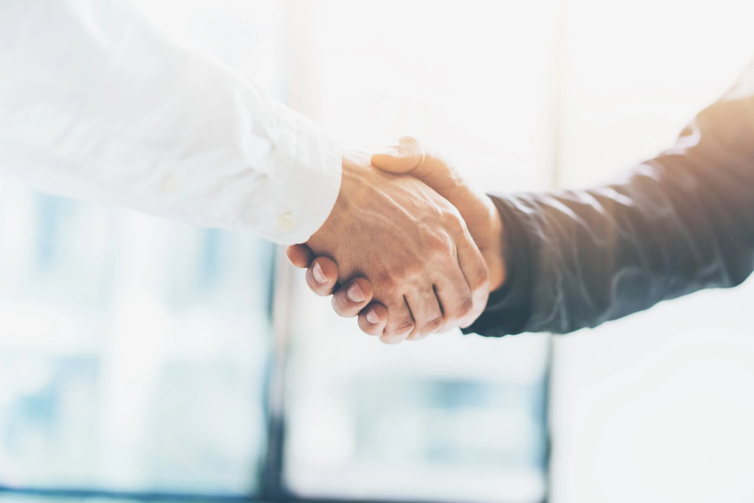 Close-up of two people shaking hands, one wearing a white shirt and the other wearing a black suit, in a professional setting.