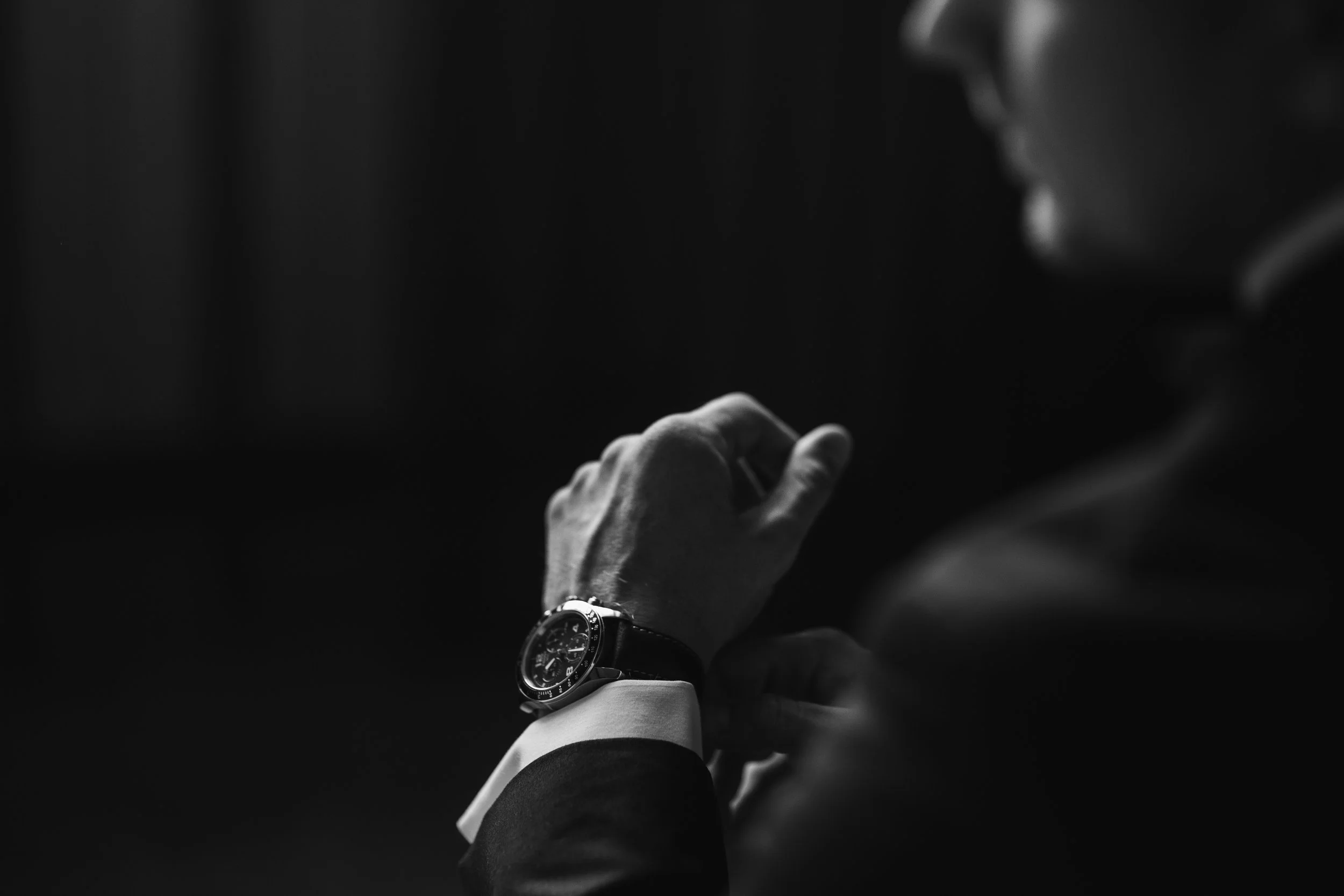 Black and white photo of a man adjusting his watch, wearing a suit, in a dimly lit environment.