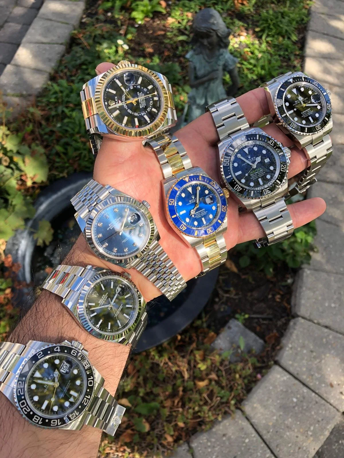 A person's arm with multiple luxury watches, including Rolex and other brands, with a garden and sidewalk in the background.