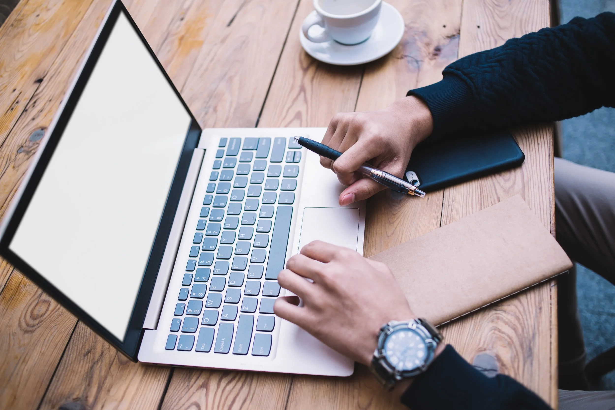 Person working on a silver laptop with a large touchpad, sitting at a wooden table, holding a pen, with a black notebook and a cup of coffee nearby.