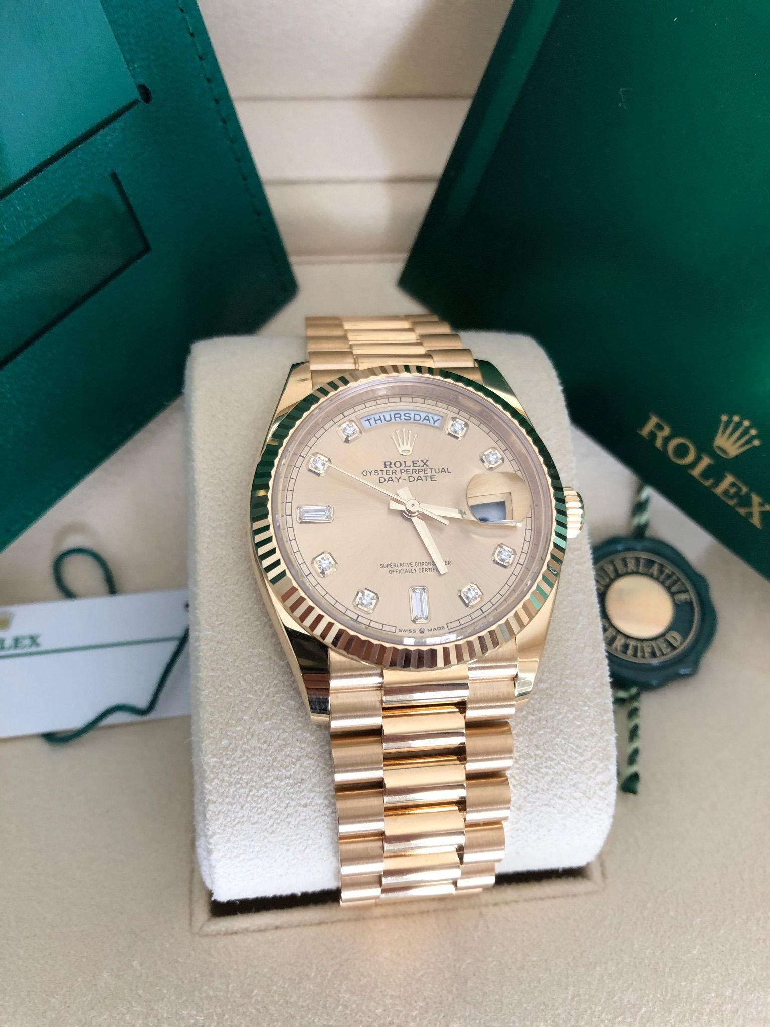 A Rolex Oyster Perpetual Day-Date watch with a gold band and diamond markers, displayed in its box with green Rolex packaging.