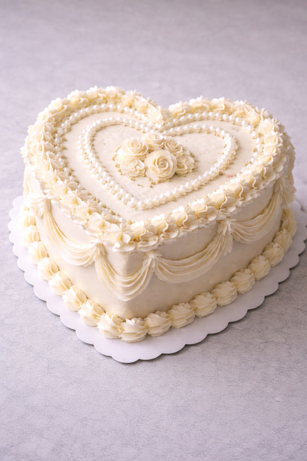 A heart-shaped white wedding cake decorated with white roses, pearl-like beads, and piped cream details, sitting on a white scalloped cake board.