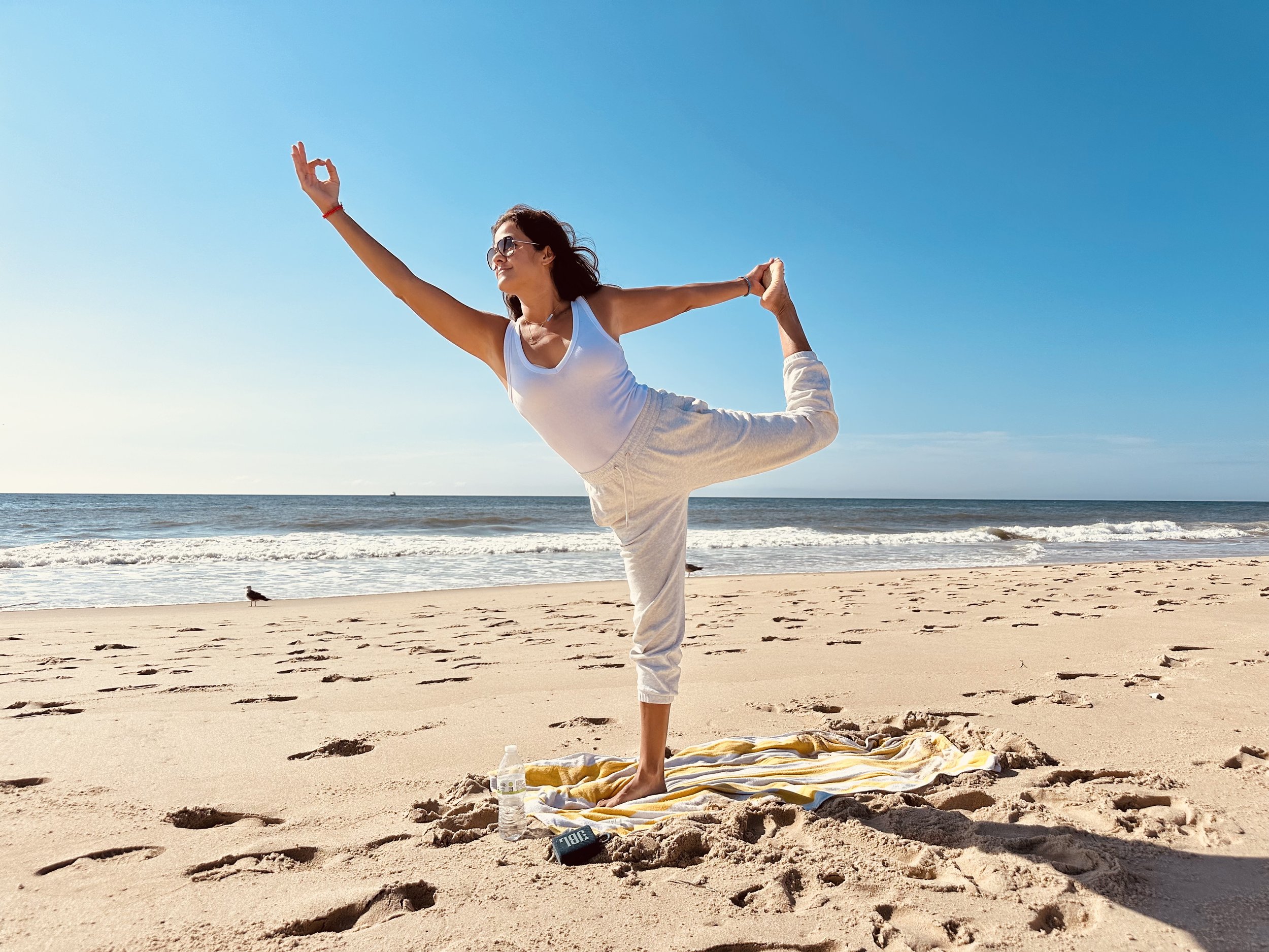 A woman practicing yoga on the beach during daytime. She is in the Natarajasana pose, balancing on one leg, with the other leg pulled back and held by her hand. She is wearing sunglasses, a white tank top, and light-colored pants. There is a towel and water bottle on the sand nearby.