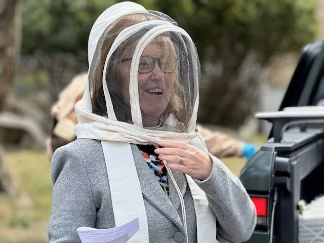 Woman in a gray blazer wearing a beekeeping suit with a veil, smiling outdoors.