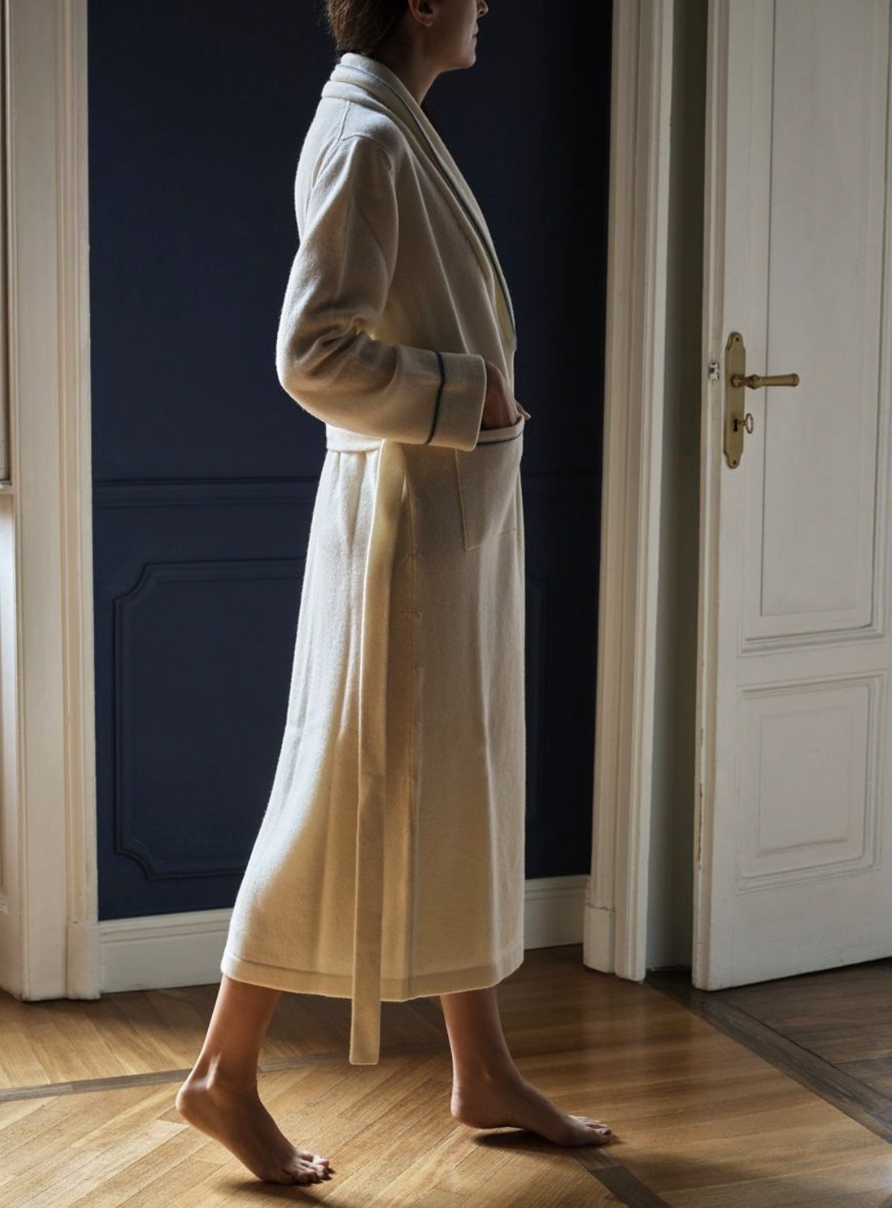 A woman standing barefoot on wooden floor in a beige bathrobe near a partially open white door of a room with dark blue walls.