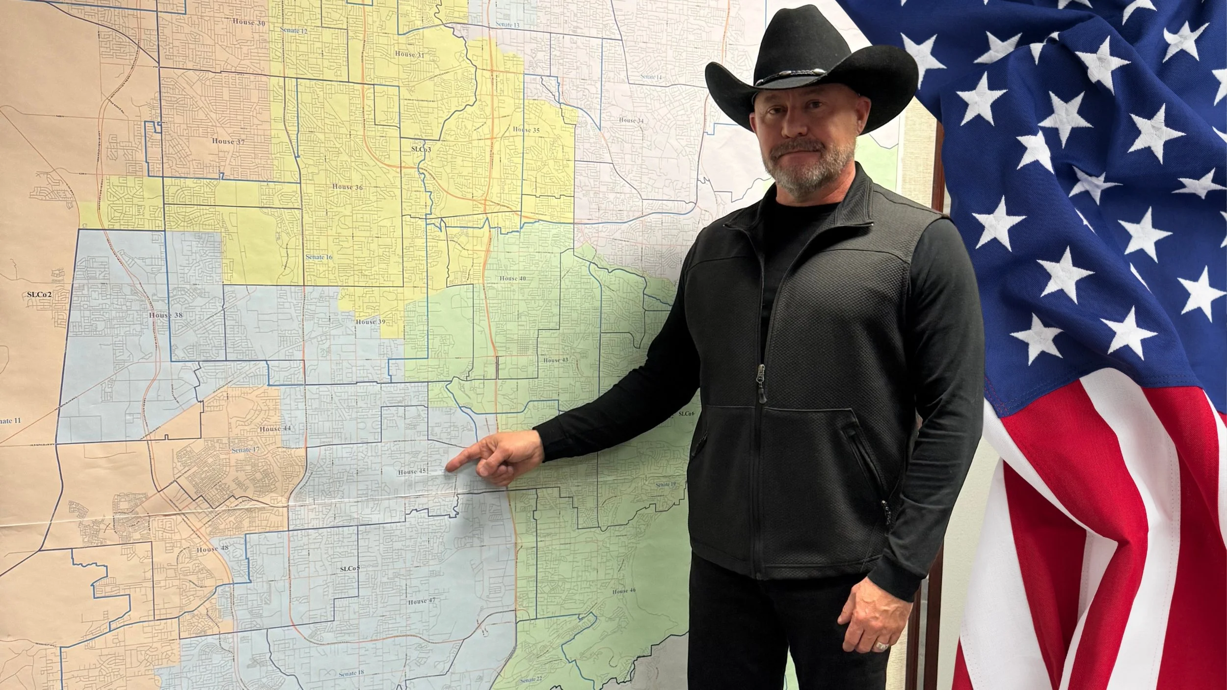 A man wearing a black cowboy hat and black vest standing in front of a map and pointing at it, with an American flag draped beside him.