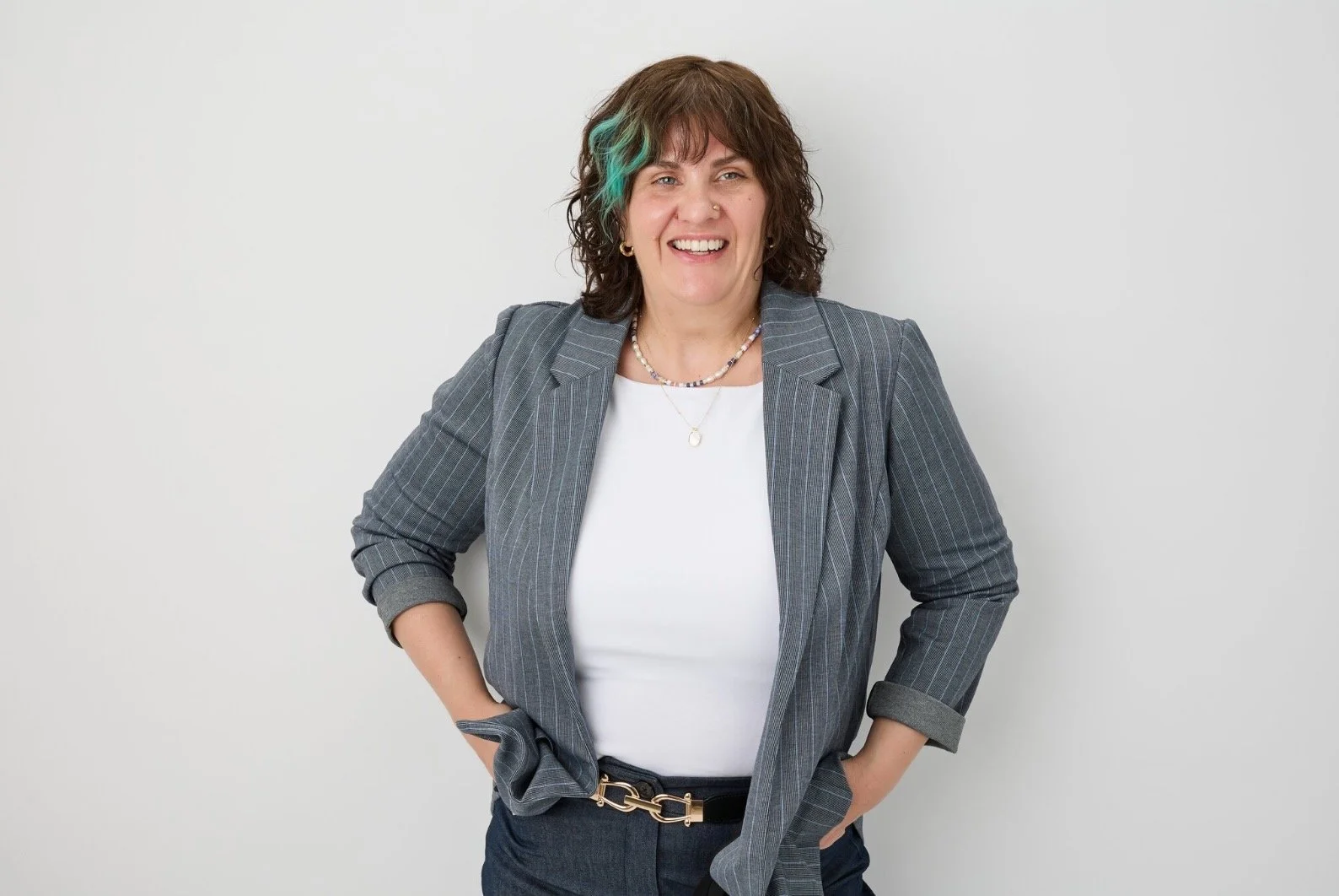 A woman with curly brown hair and teal highlights, smiling, wearing a gray pinstripe blazer, white top, and dark pants, standing against a plain white wall.
