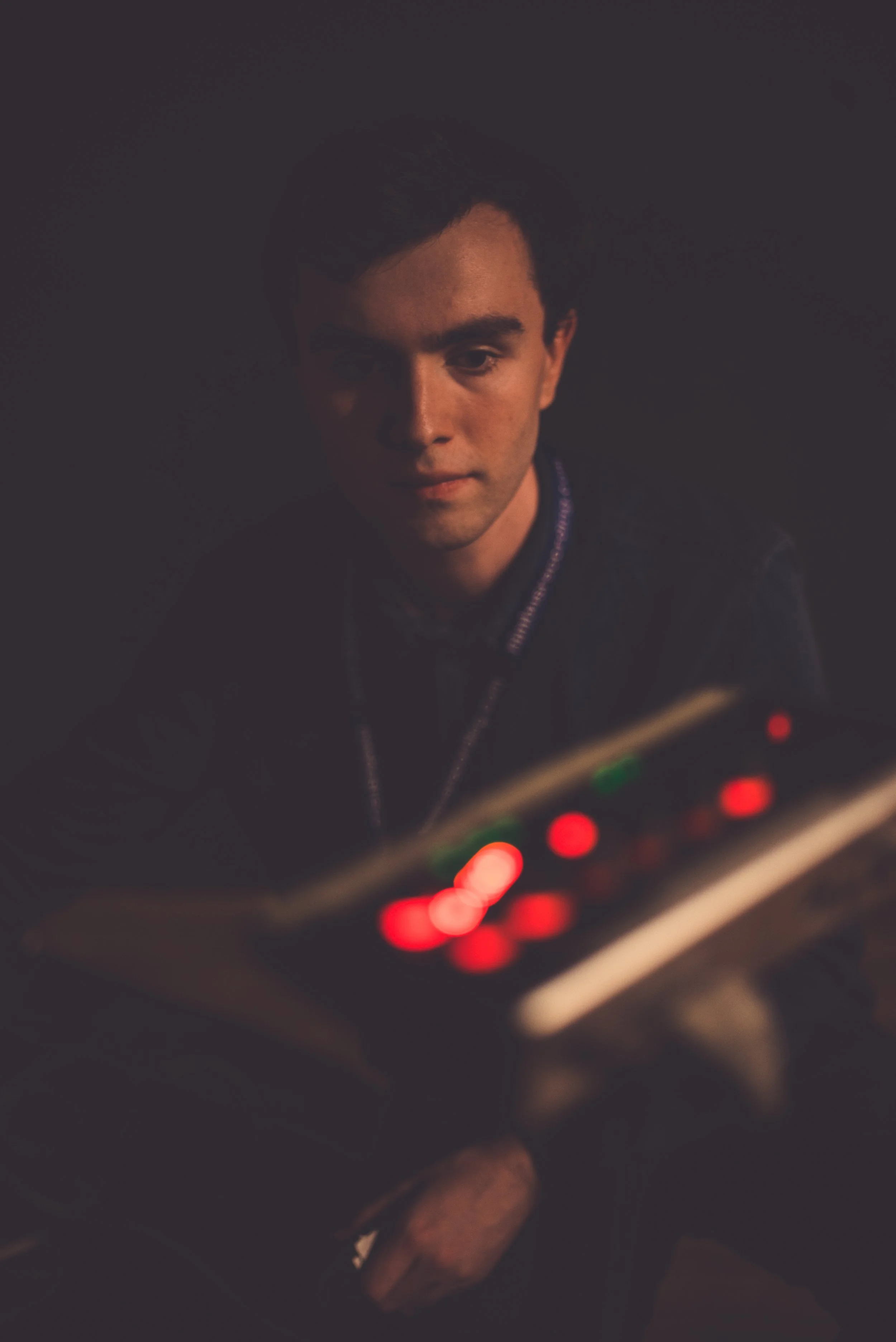 A young man with dark hair and a slight smile, sitting in dim lighting, holding and looking at a remote control with red and green buttons.