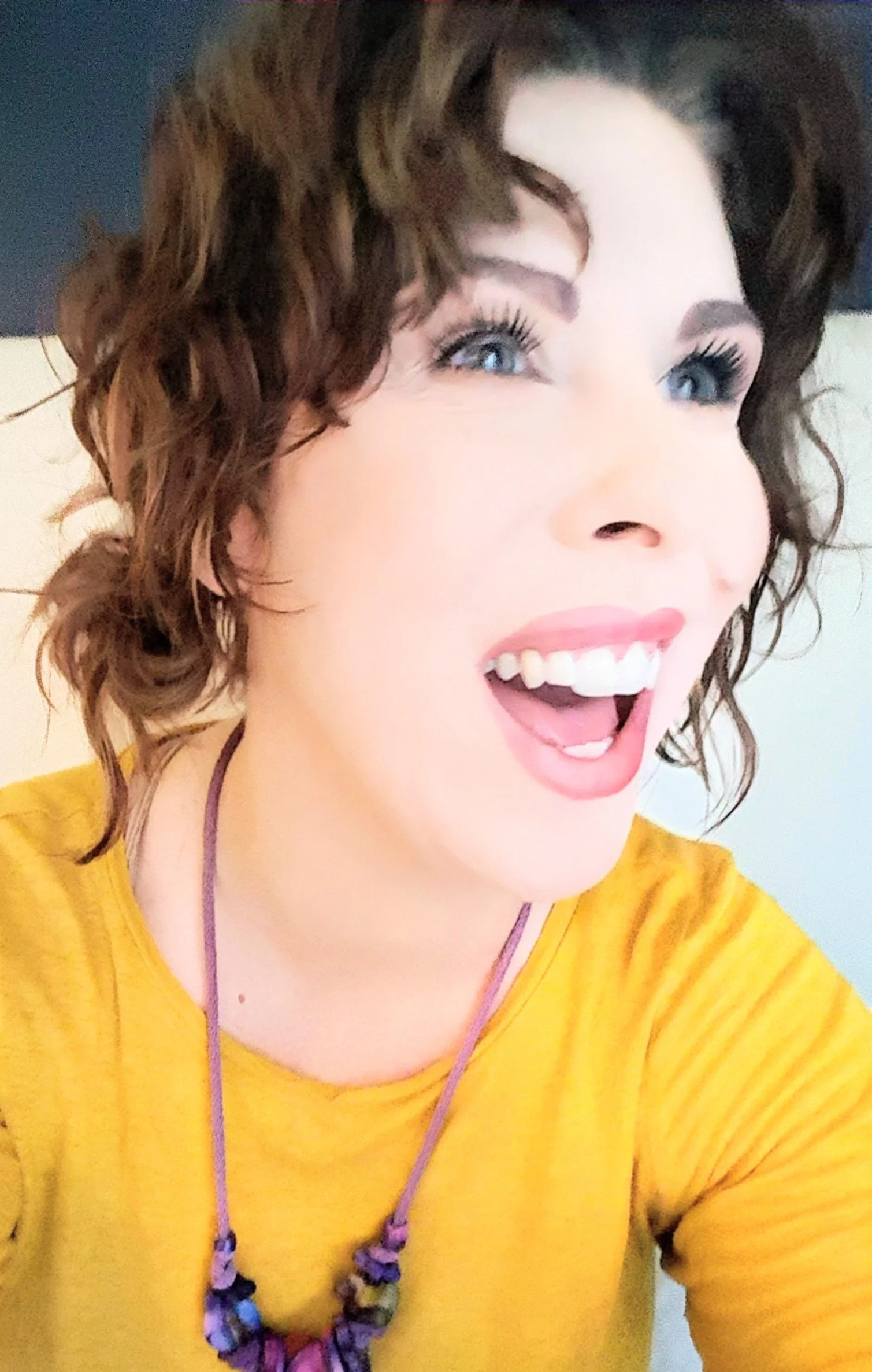 A woman with short, curly brown hair and bright blue eyes is smiling widely with her mouth open, wearing a yellow top and a purple beaded necklace.