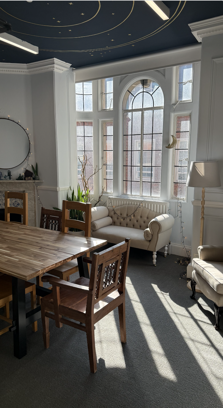A sunlit living room with large windows, a white tufted loveseat, a beige armchair, a tall floor lamp, and a wooden dining table with chairs. The ceiling is dark blue with gold star and celestial designs, and decorative moon and star hanging accents are near the windows. String lights are draped along the wall.