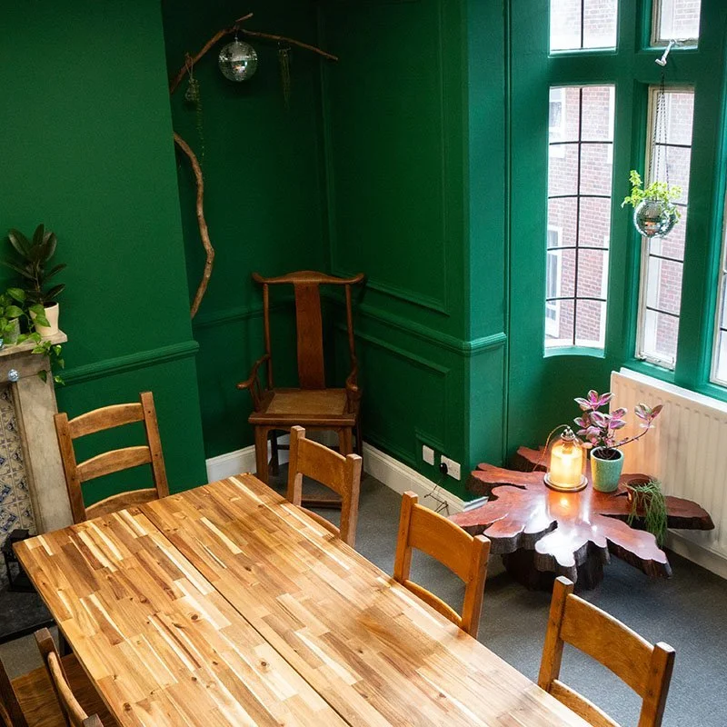 A cozy dining area with a wooden table and six chairs, green walls with decorative paneling, a corner with a wooden chair, and a window sill with plants and a lit candle.