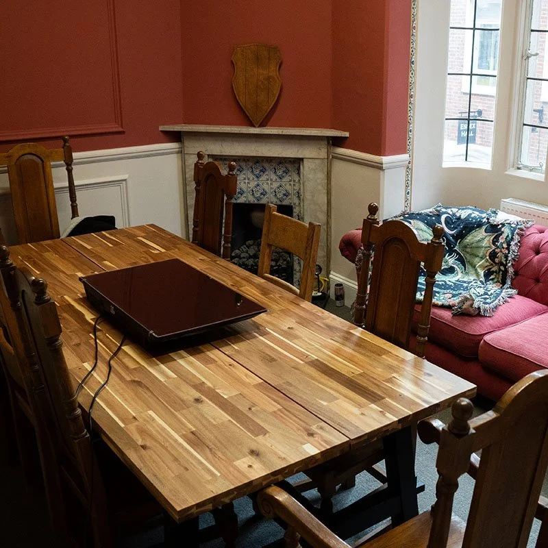 A wooden dining table with a closed laptop on top, surrounded by wooden chairs, in a dining room with pink and white walls, a fireplace with a decorative wooden shield above it, a pink sofa with a blanket, and a window letting in natural light.