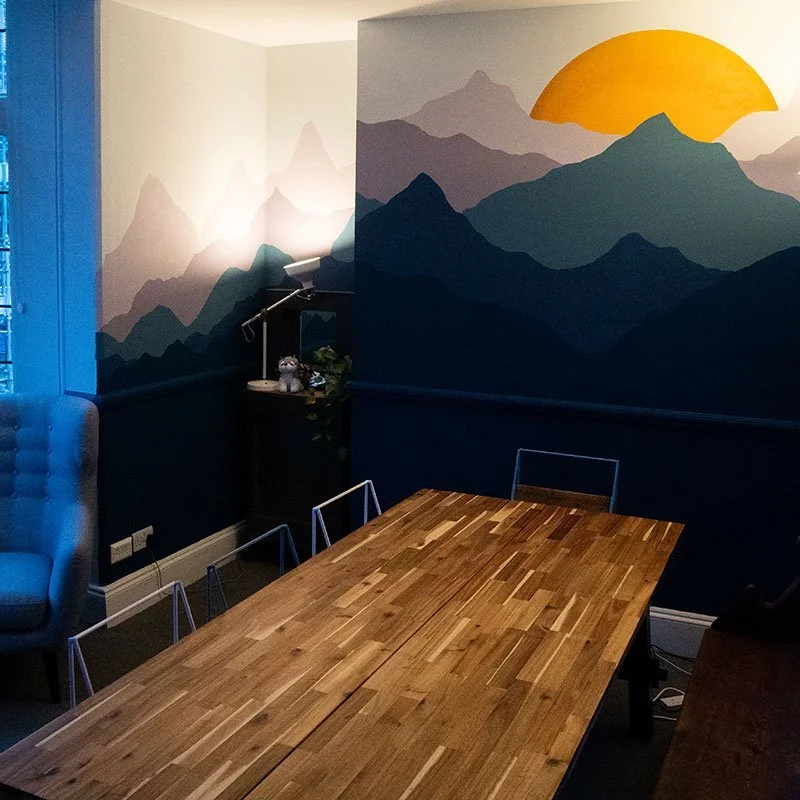 A mural of mountain silhouettes with a yellow sun on the wall behind a wooden table in a room.