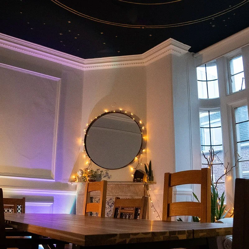 A dining room with a round mirror decorated with string lights on a beige wall, a large bay window with a view outside, and a ceiling with a starry sky mural featuring gold and white rings.
