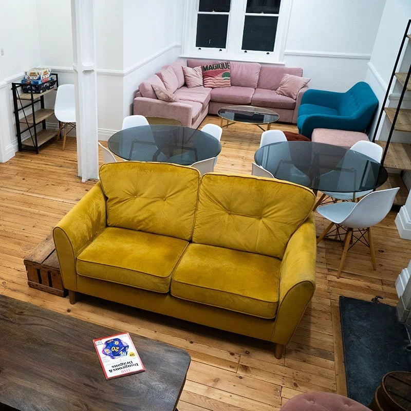 A cozy living room with a yellow velvet sofa, white and acrylic chairs around glass-top tables, a pink sectional sofa with cushions, a blue armchair, a wooden staircase, and hardwood floors.