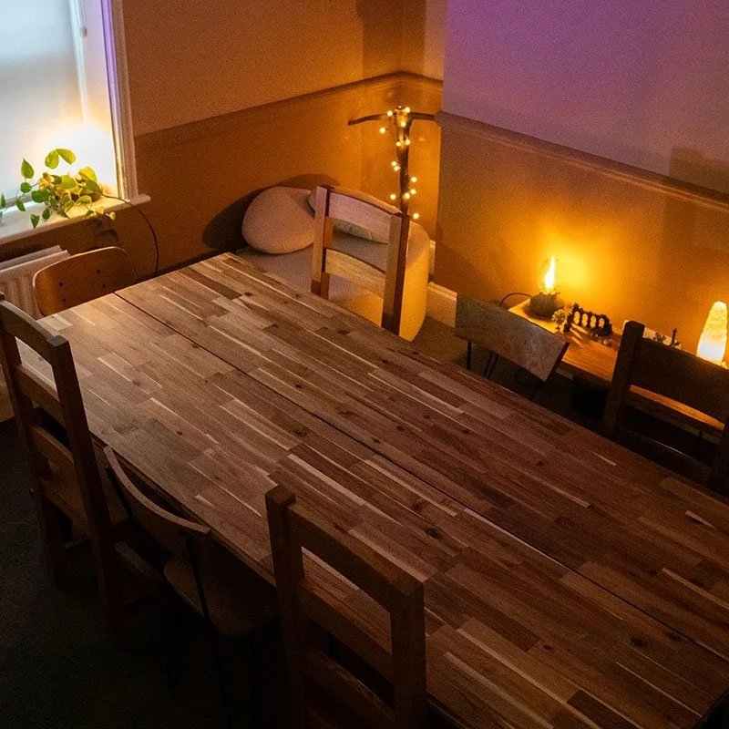 A wooden dining table in a cozy room with string lights, a potted plant by the window, and warm lighting on a side table.